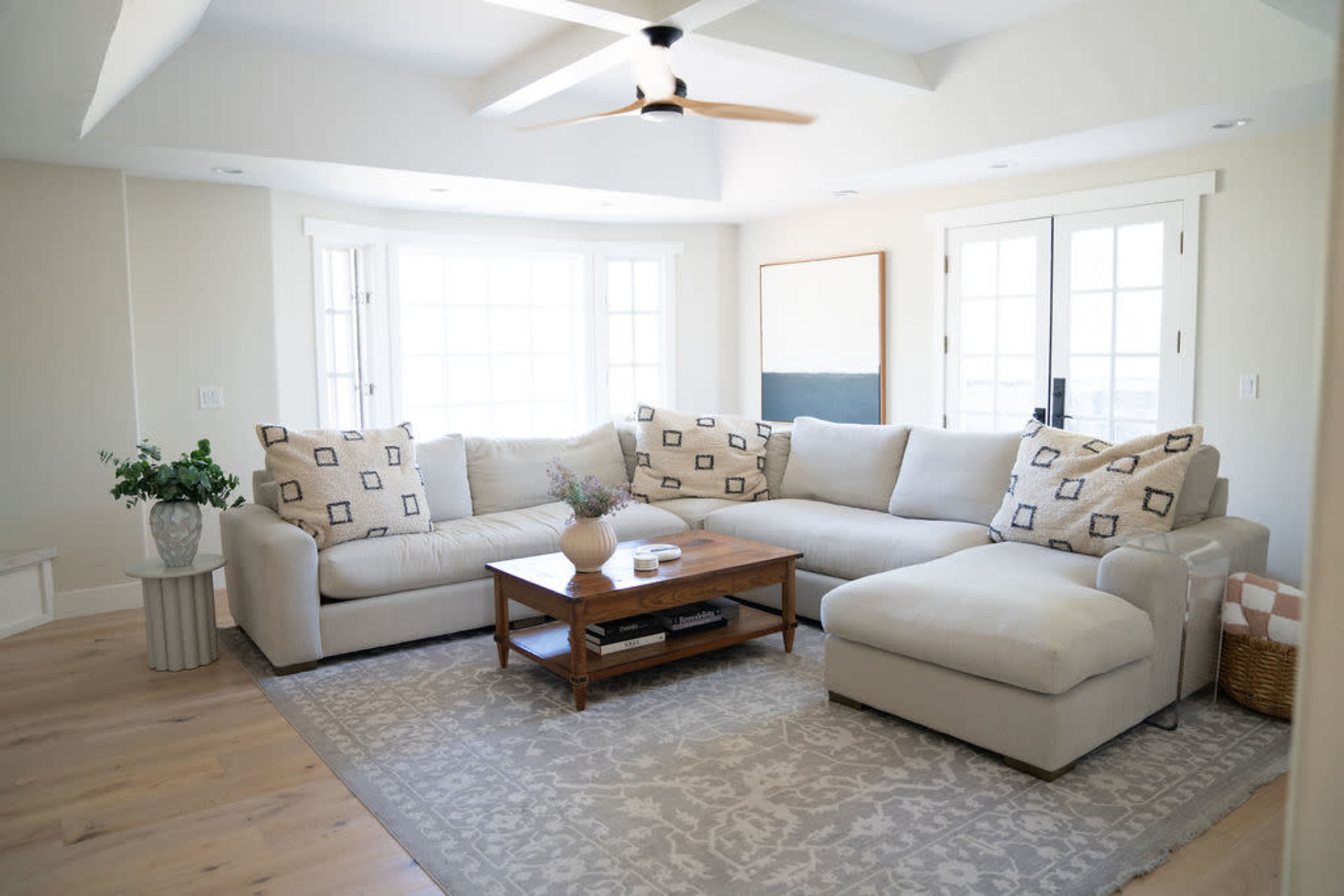 A light-filled living room featuring a large sectional sofa, a wooden coffee table, and a neutral area rug, with windows overlooking an outdoor space.