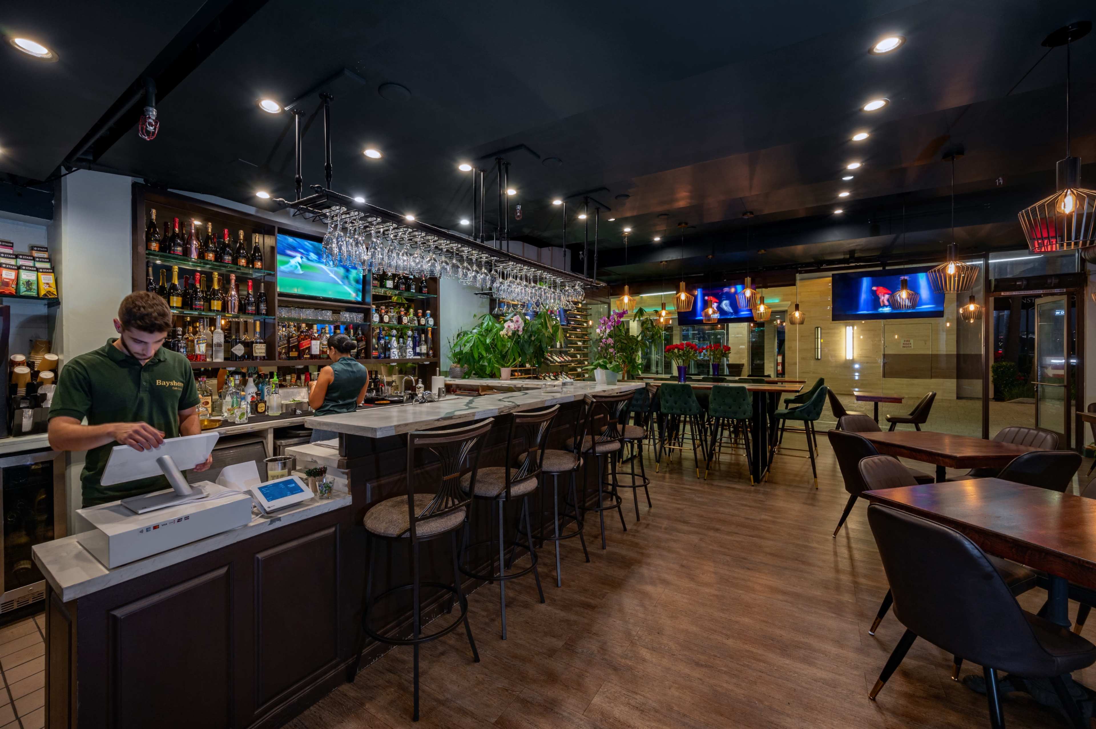 A modern bar interior features a counter with a cash register, shelves stocked with liquor, and television screens displaying entertainment, surrounded by tables and chairs.