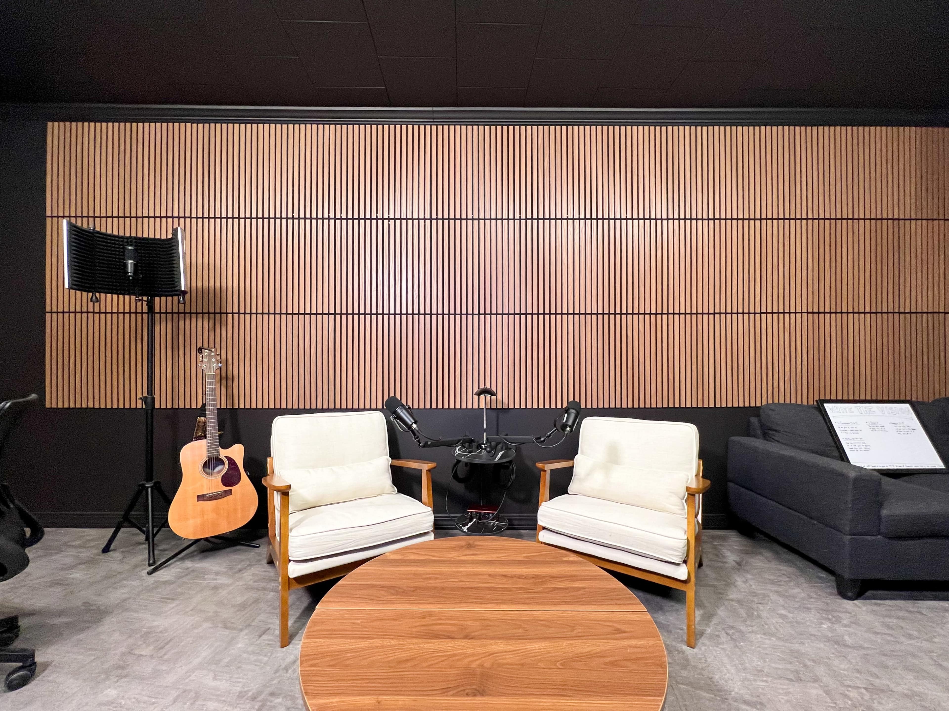 The image shows a modern interior space featuring two white armchairs facing a round wooden table, with a guitar and an audio stand in the background against a wood-slatted wall.