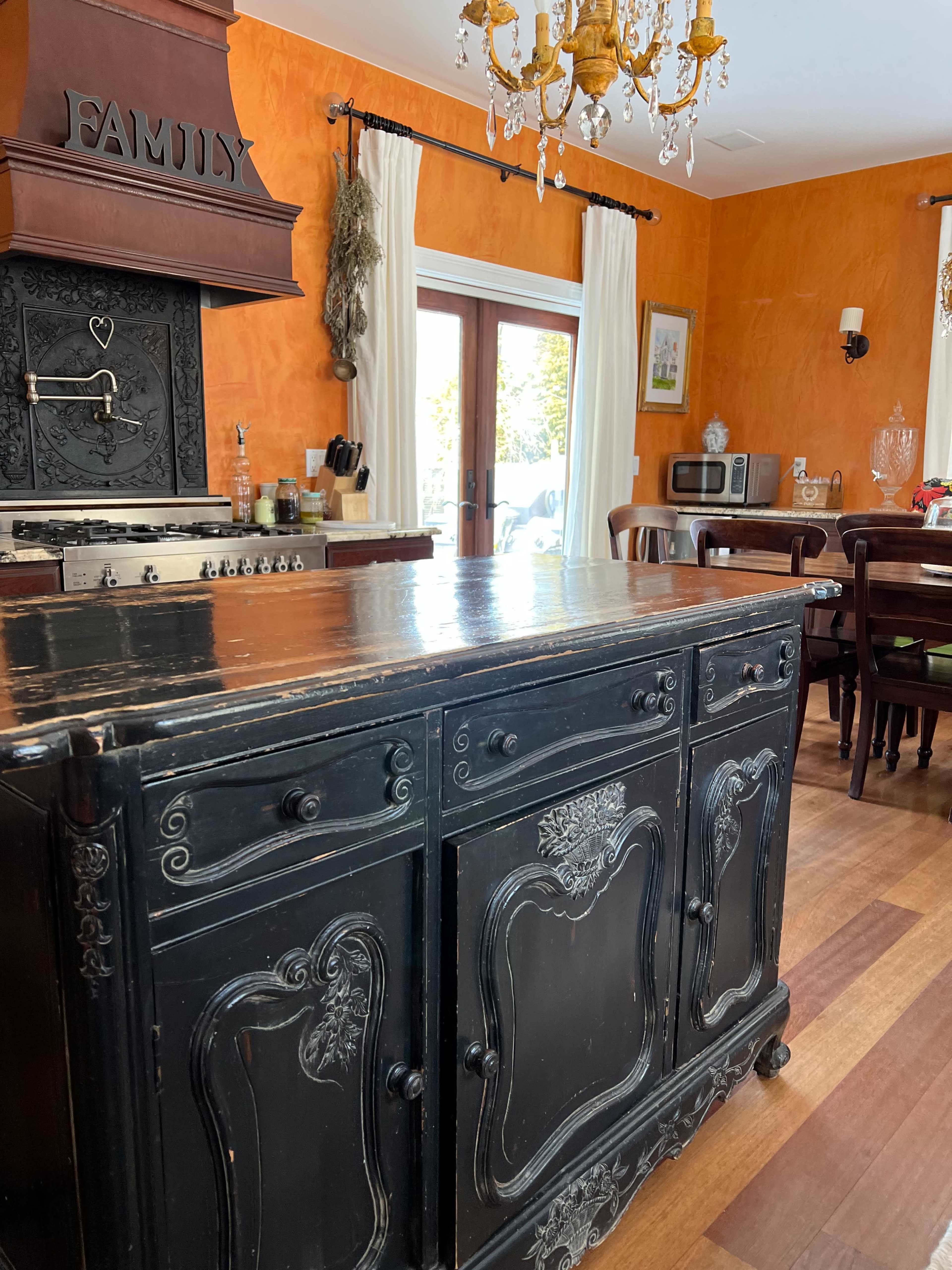 A black wooden sideboard with intricate detailing is positioned in a brightly colored kitchen featuring an orange wall and a chandelier overhead.