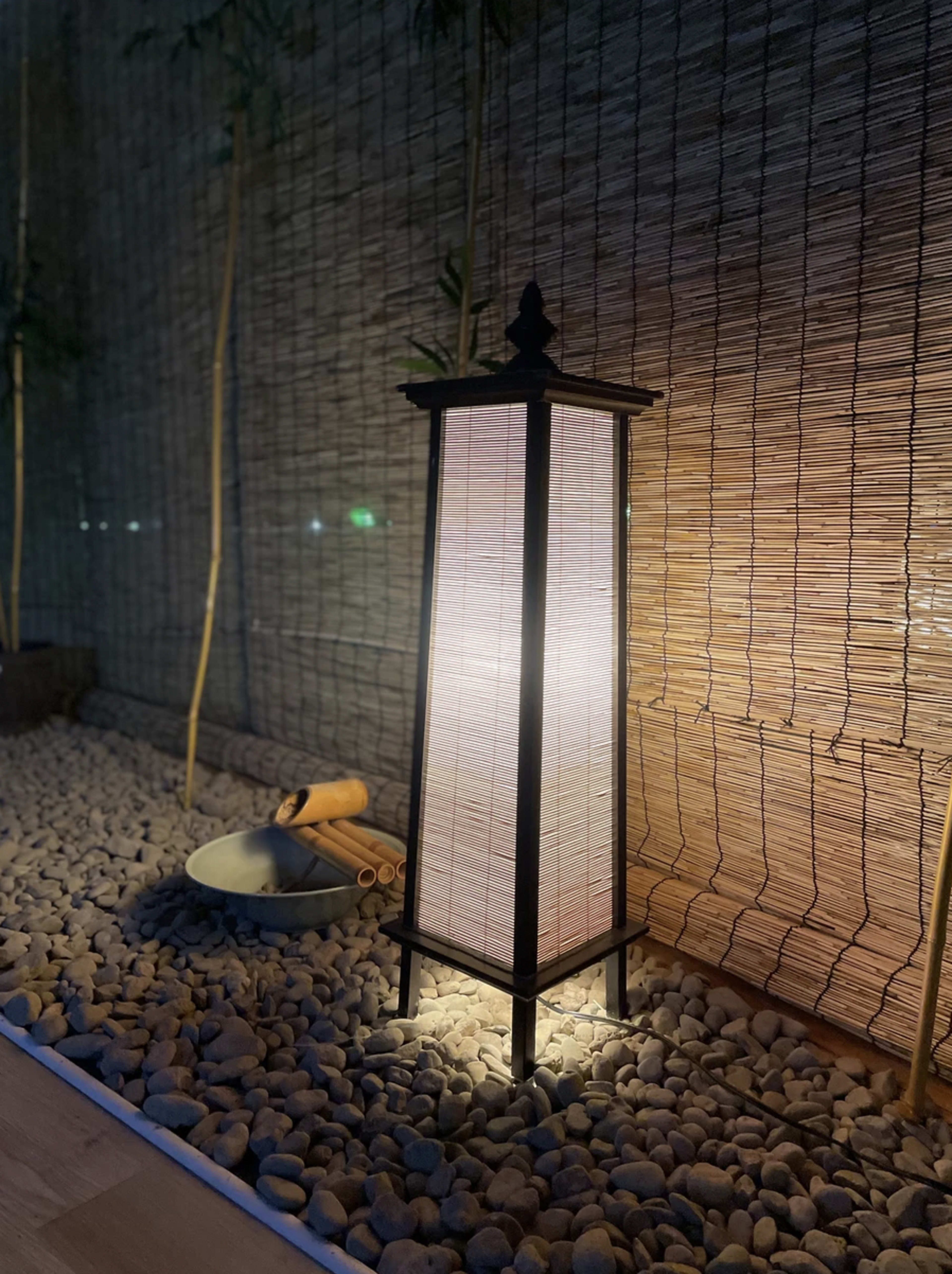 A lit lantern stands beside a bowl and bamboo on a bed of smooth pebbles against a textured wall.