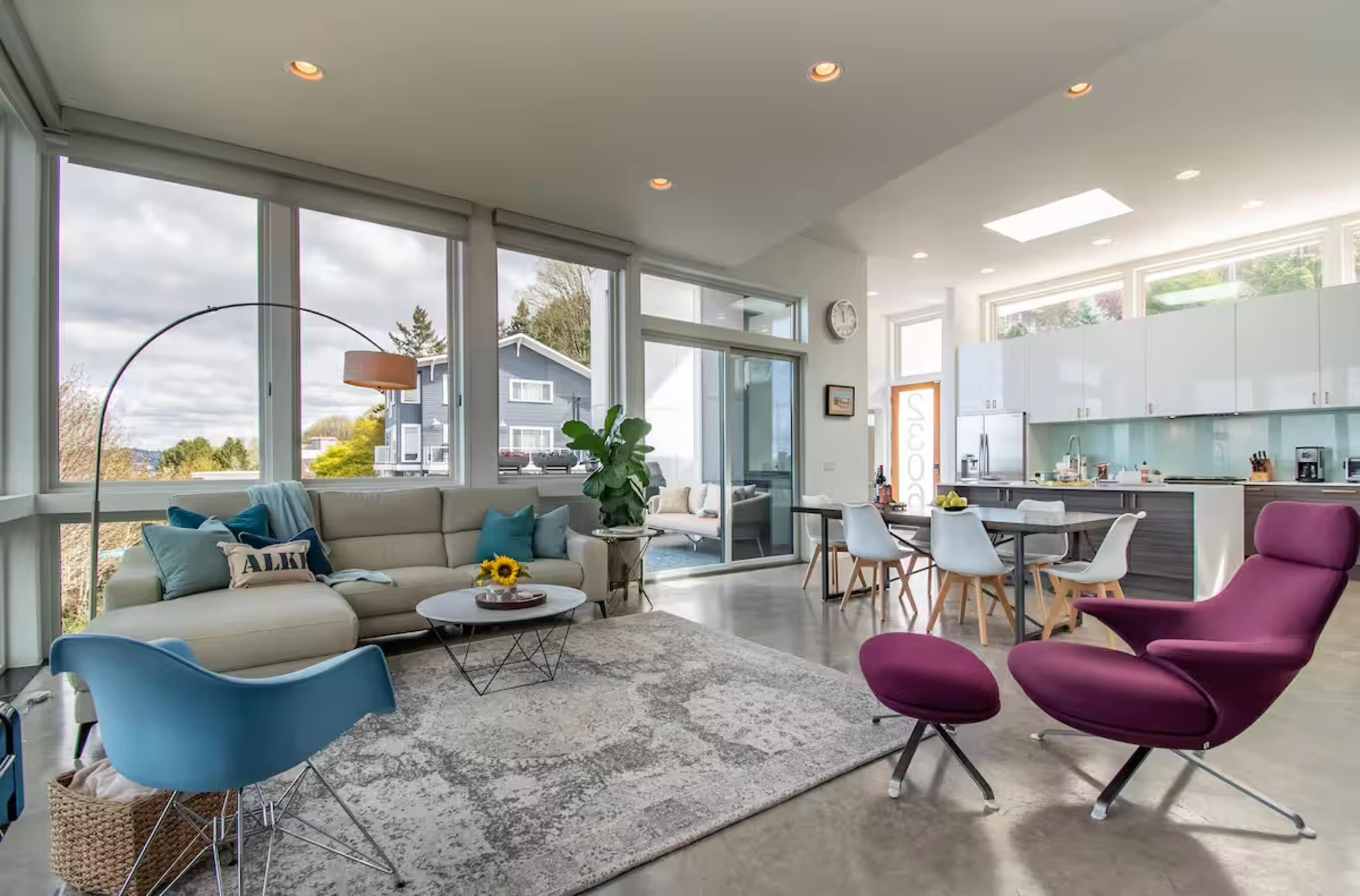 The image shows a modern living room with large windows, a light-colored sofa, a round coffee table, a dining area with white chairs, and a vibrant purple lounge chair.