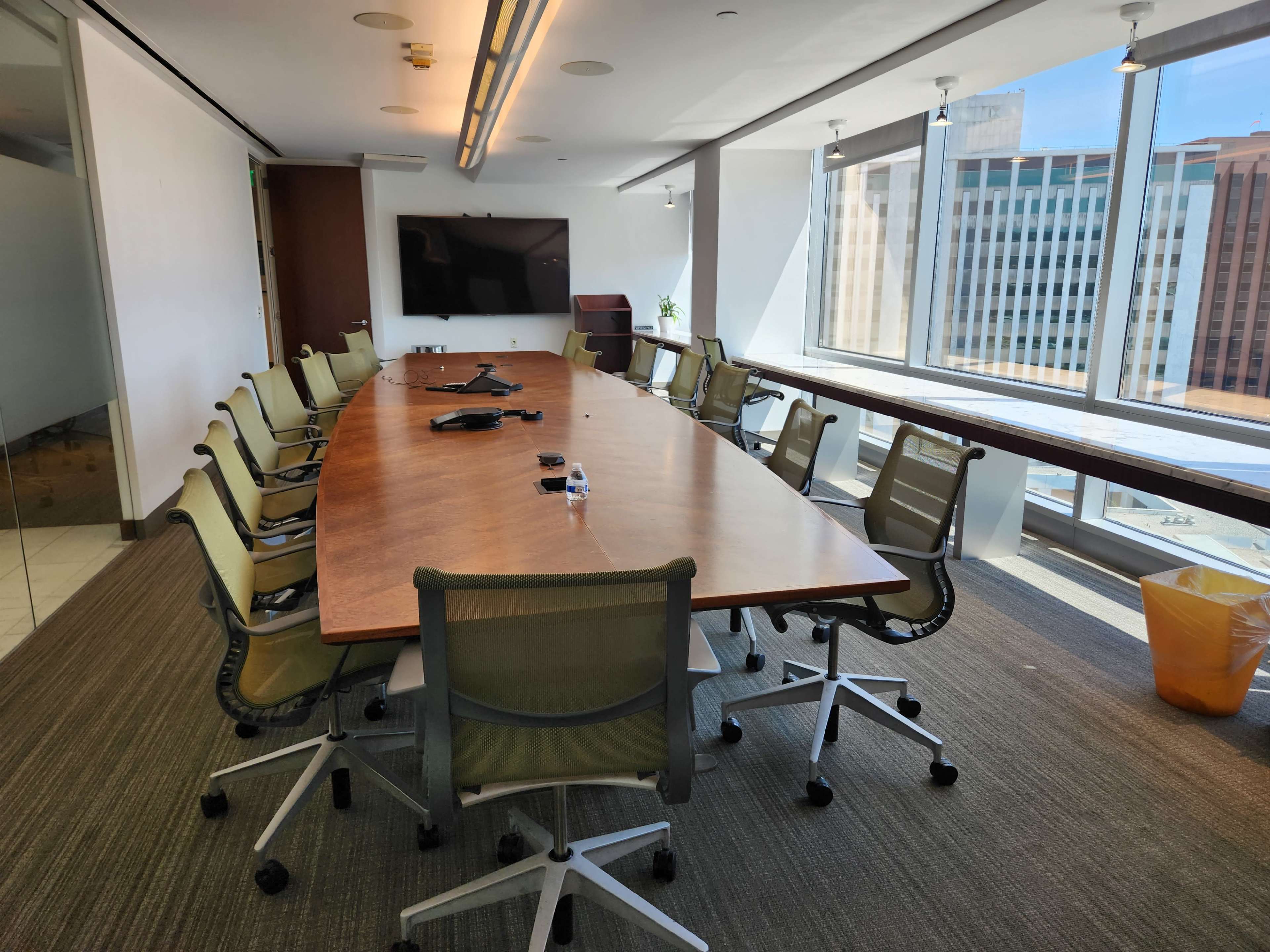 A large conference room features a long wooden table surrounded by ergonomic chairs, with windows offering a view of a city skyline.