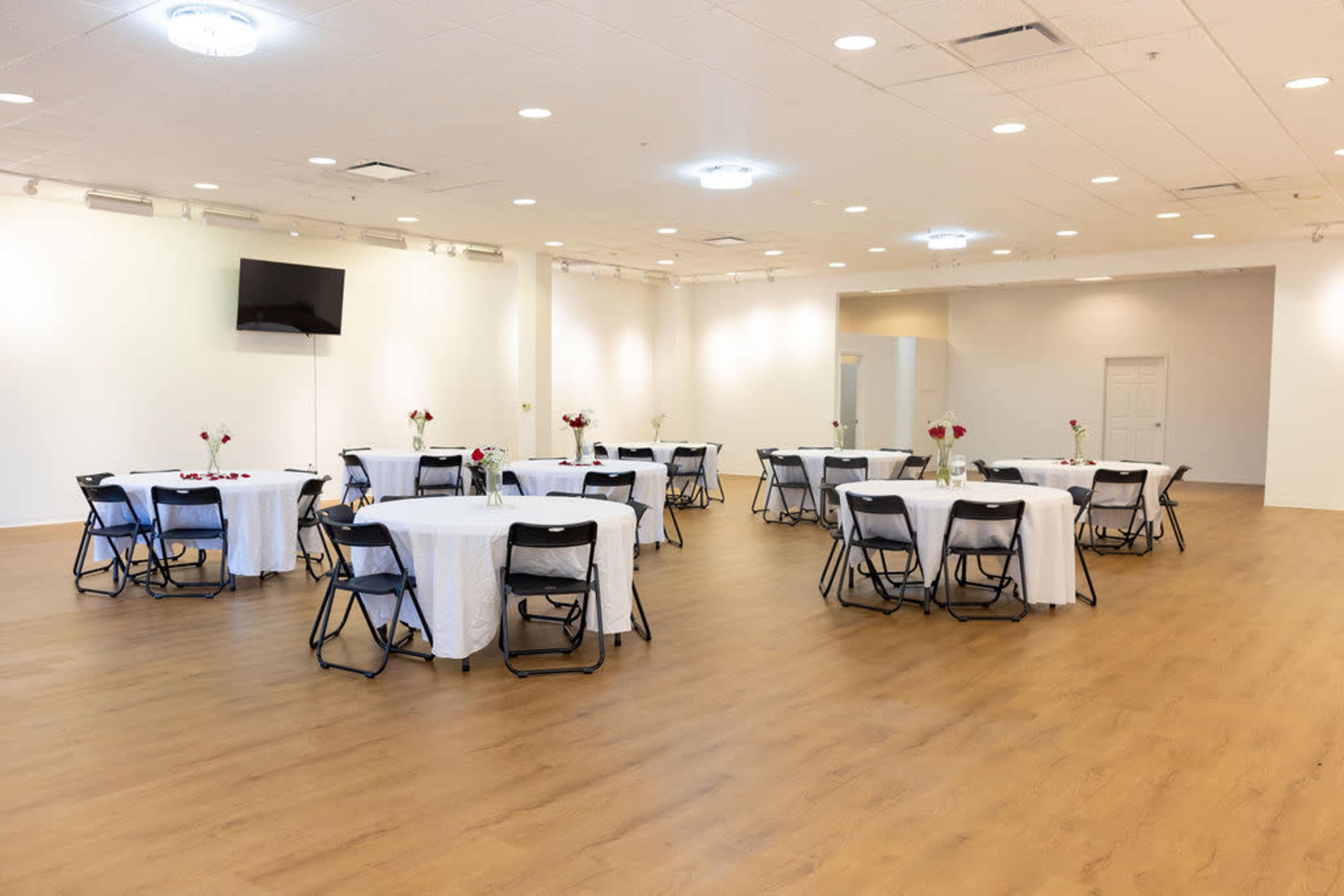 The image shows a spacious event hall with several round tables set with white tablecloths and chairs arranged neatly, featuring flower vases as centerpieces.