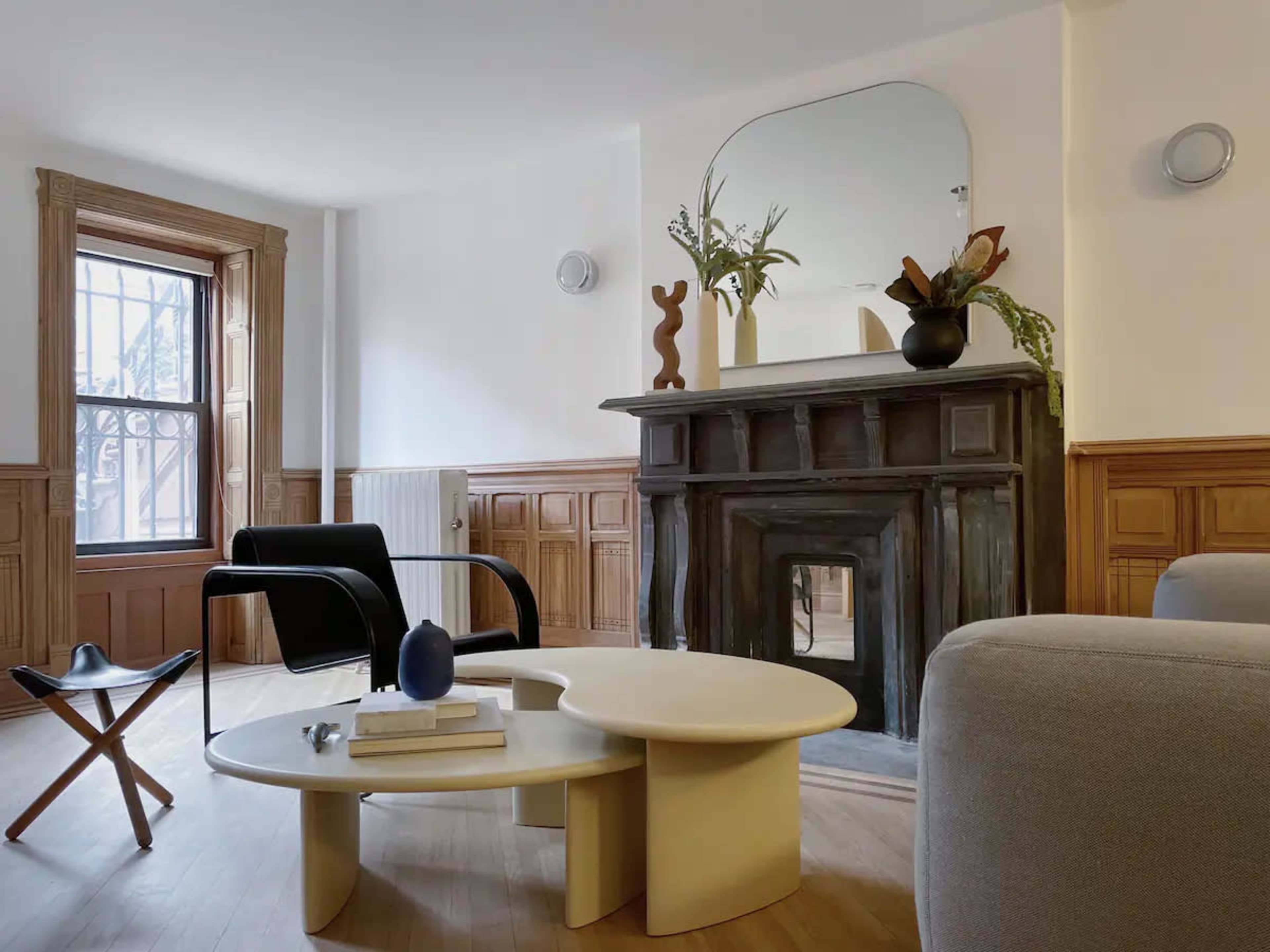The image shows a modern living room featuring a black armchair, a light-colored coffee table, a decorative fireplace, and a large mirror above it.
