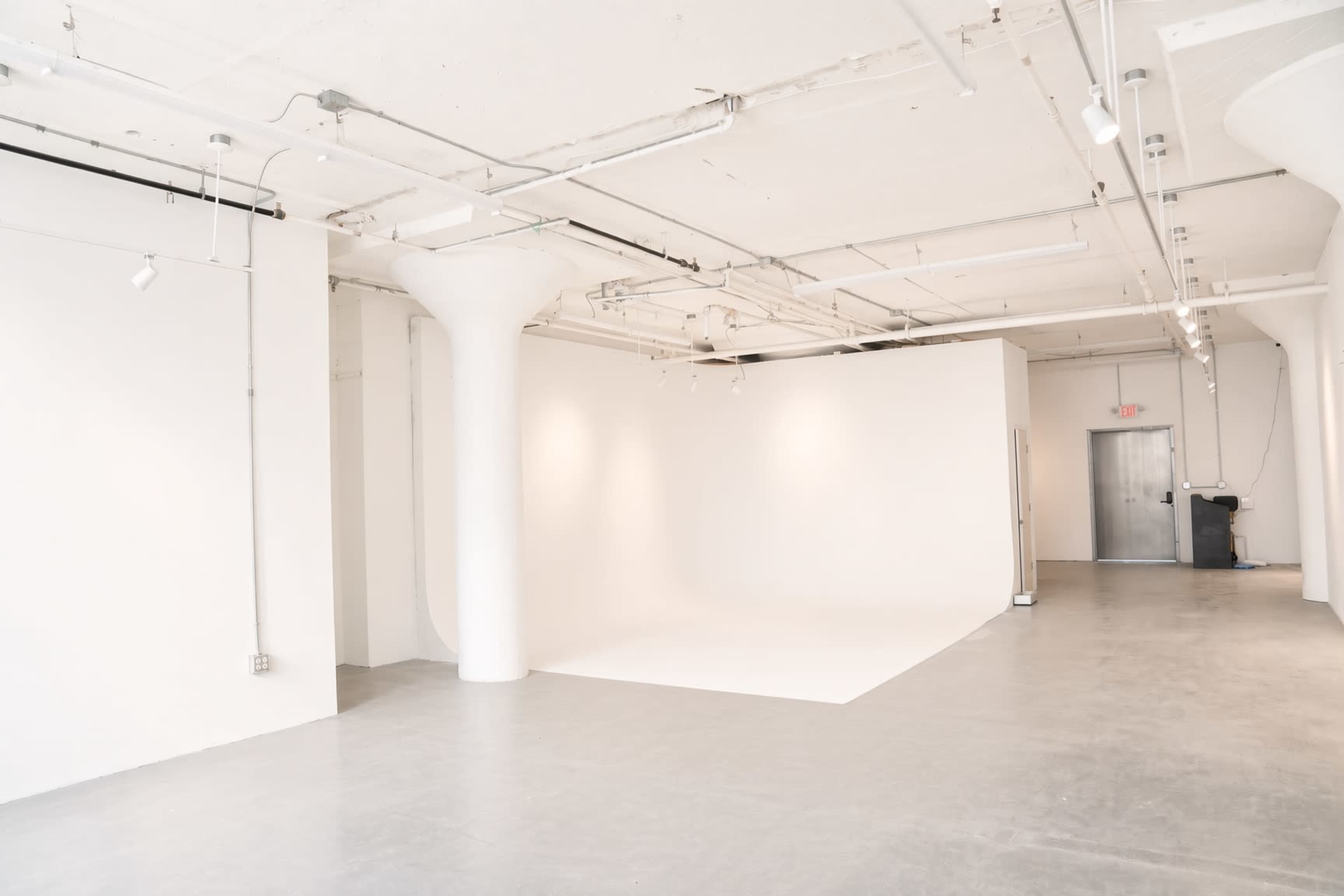 The image shows a spacious, minimalist room with white walls and a concrete floor, featuring a blank backdrop and overhead lighting.