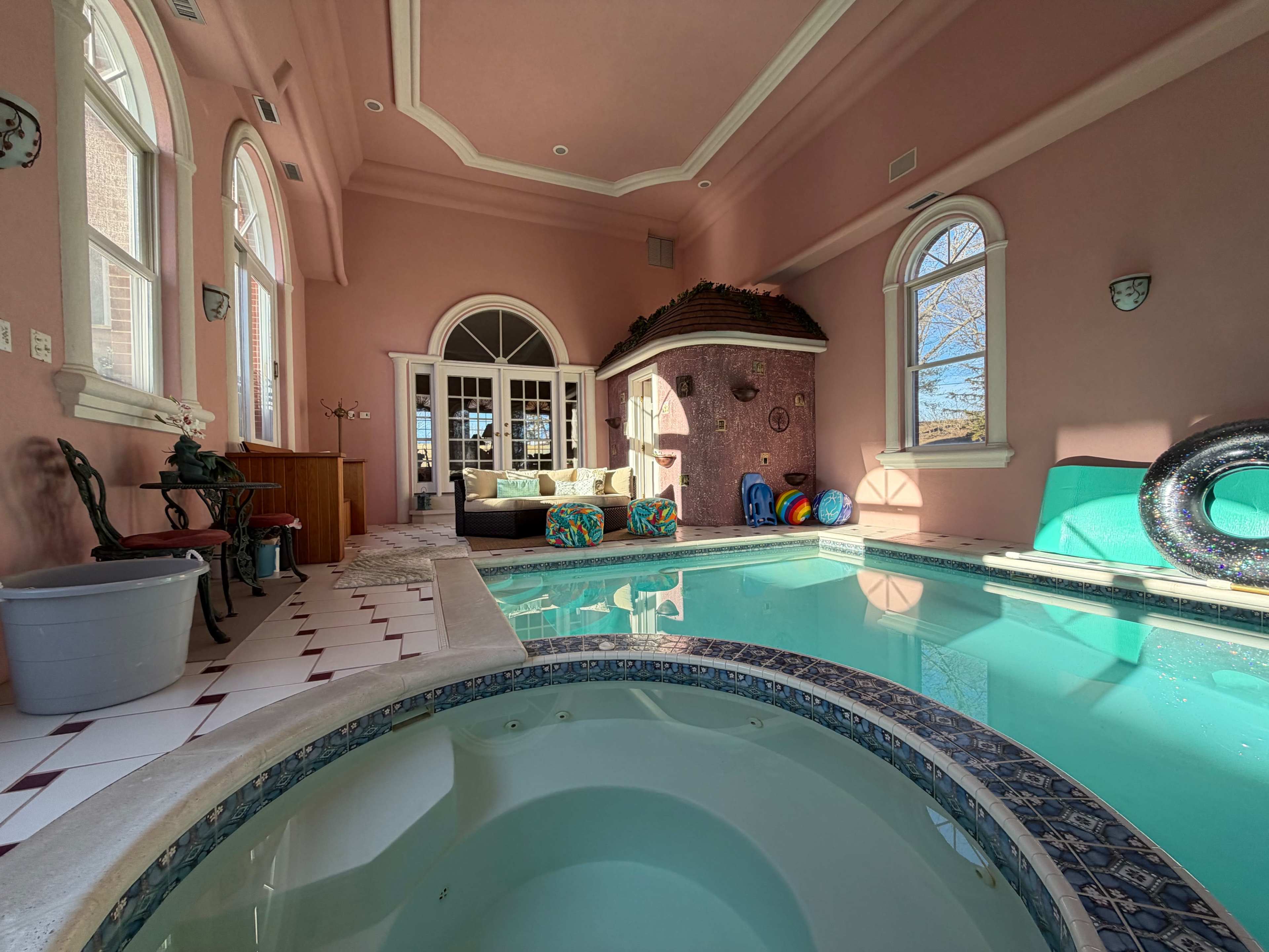 The image shows an indoor swimming pool area with pink walls, a jacuzzi, and colorful lounge furniture beside large windows.