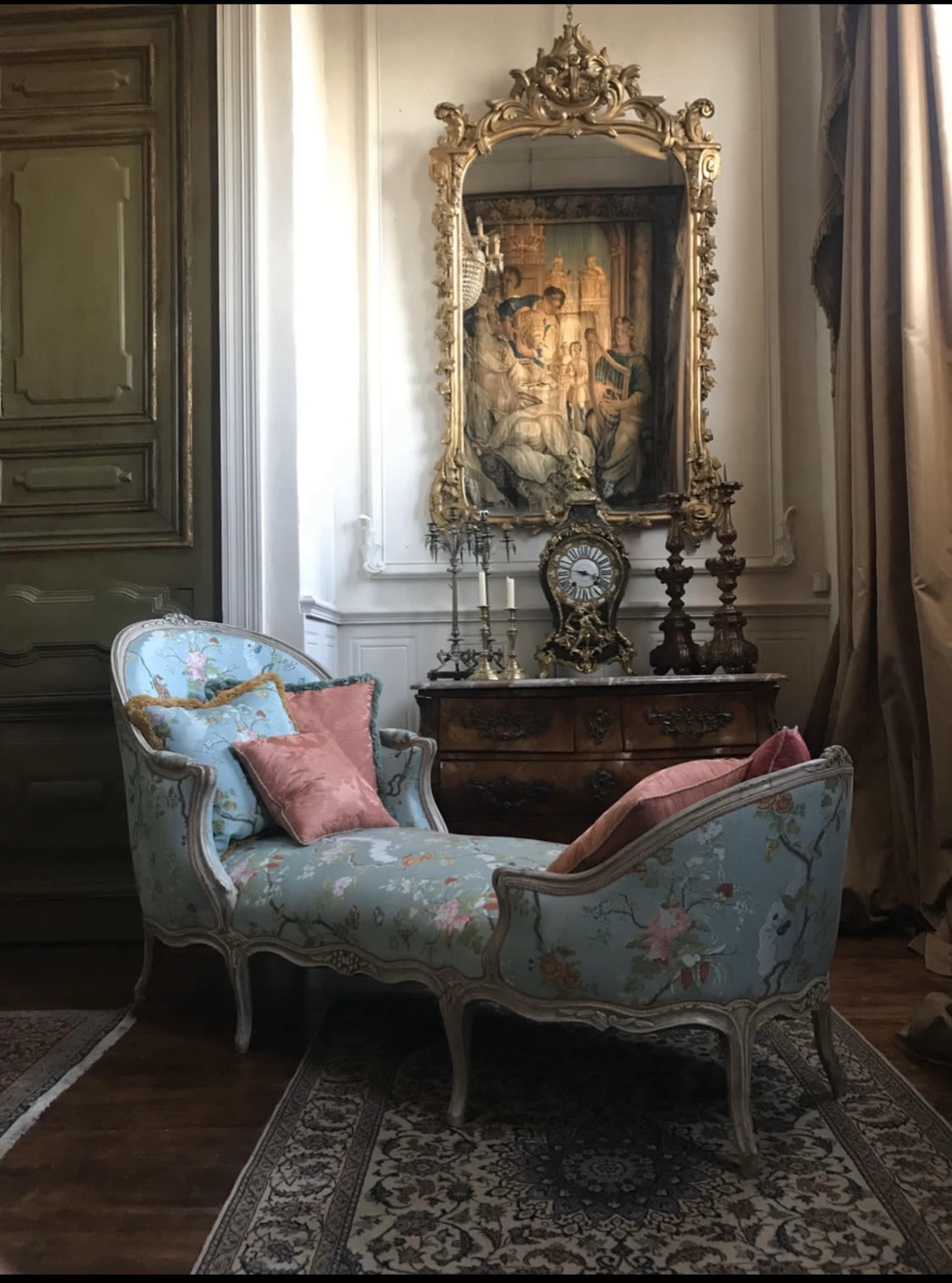 A floral-patterned chaise lounge with pink cushions is positioned near an ornate mirror and antique furniture in a well-decorated room.