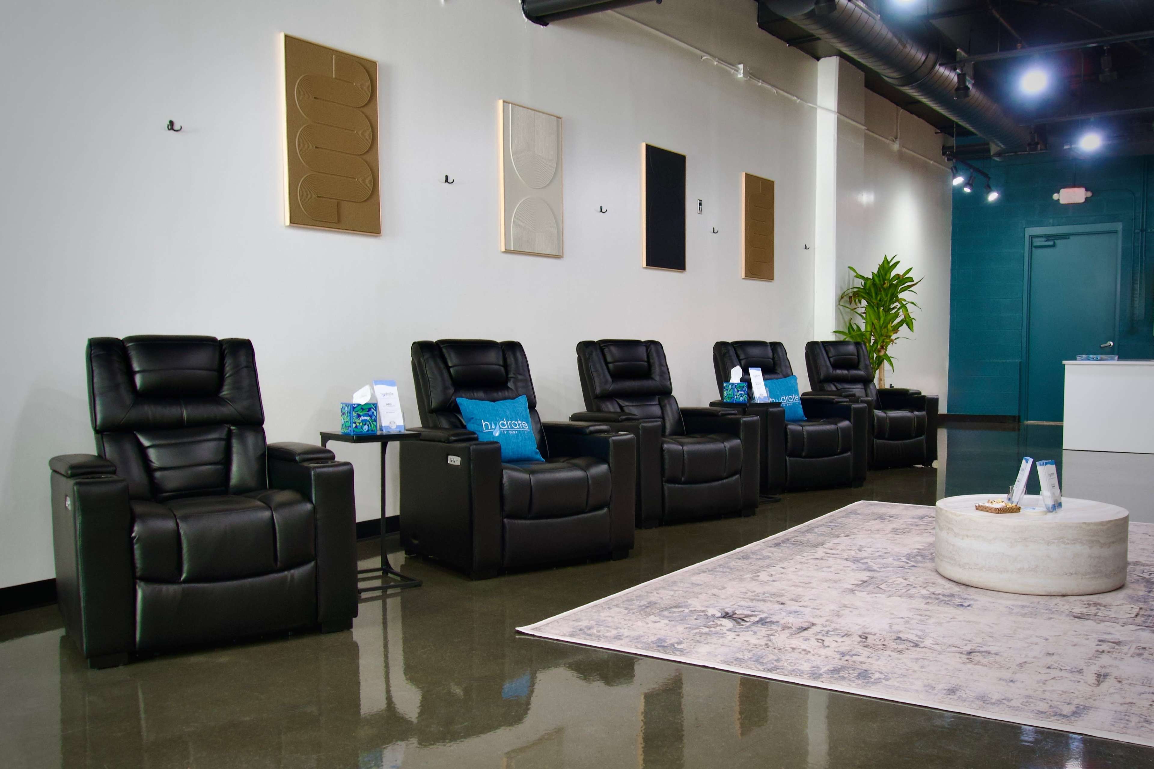 The image shows a modern waiting area with four black reclining chairs facing a round coffee table on a patterned rug, decorated with wall art in neutral tones.