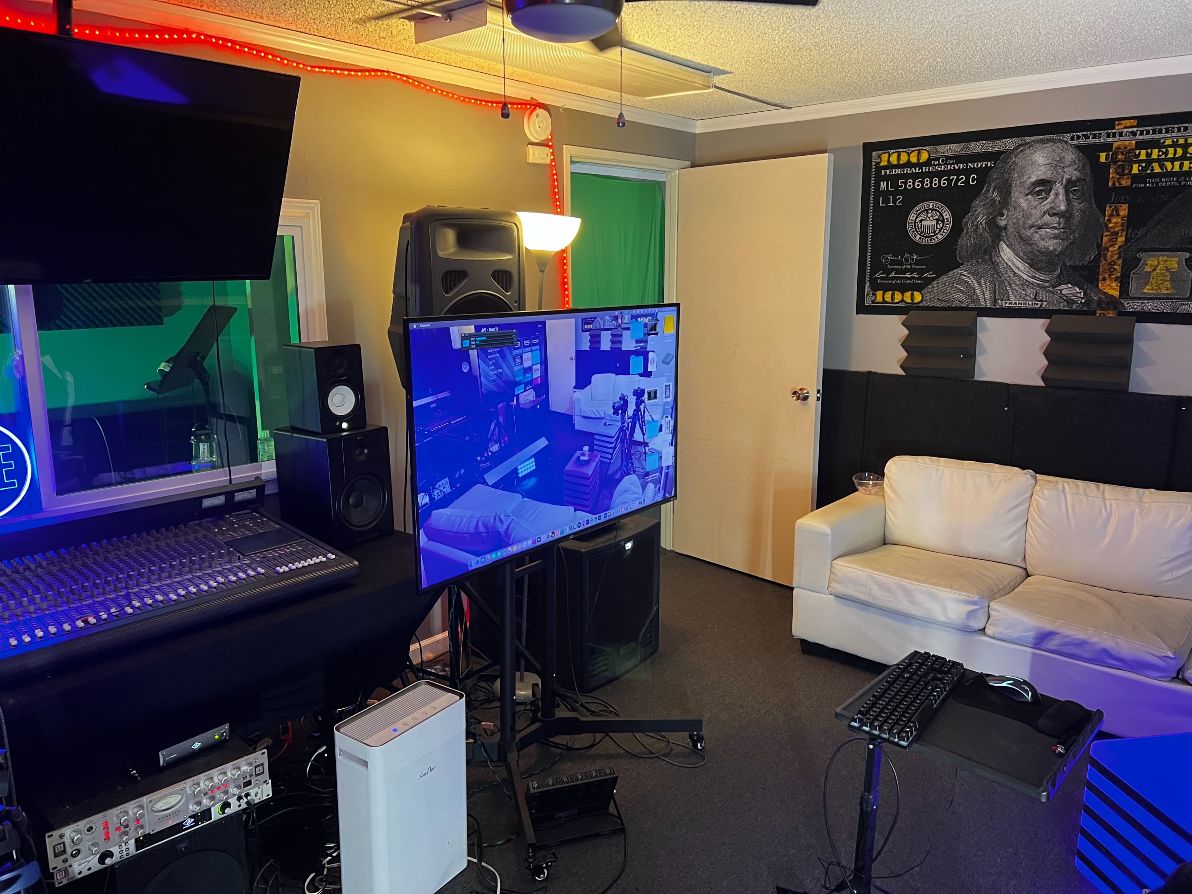 The image shows a multimedia studio setup with a mixing console, a large television displaying a gaming interface, and a sofa against the wall, all accented by colorful lighting.