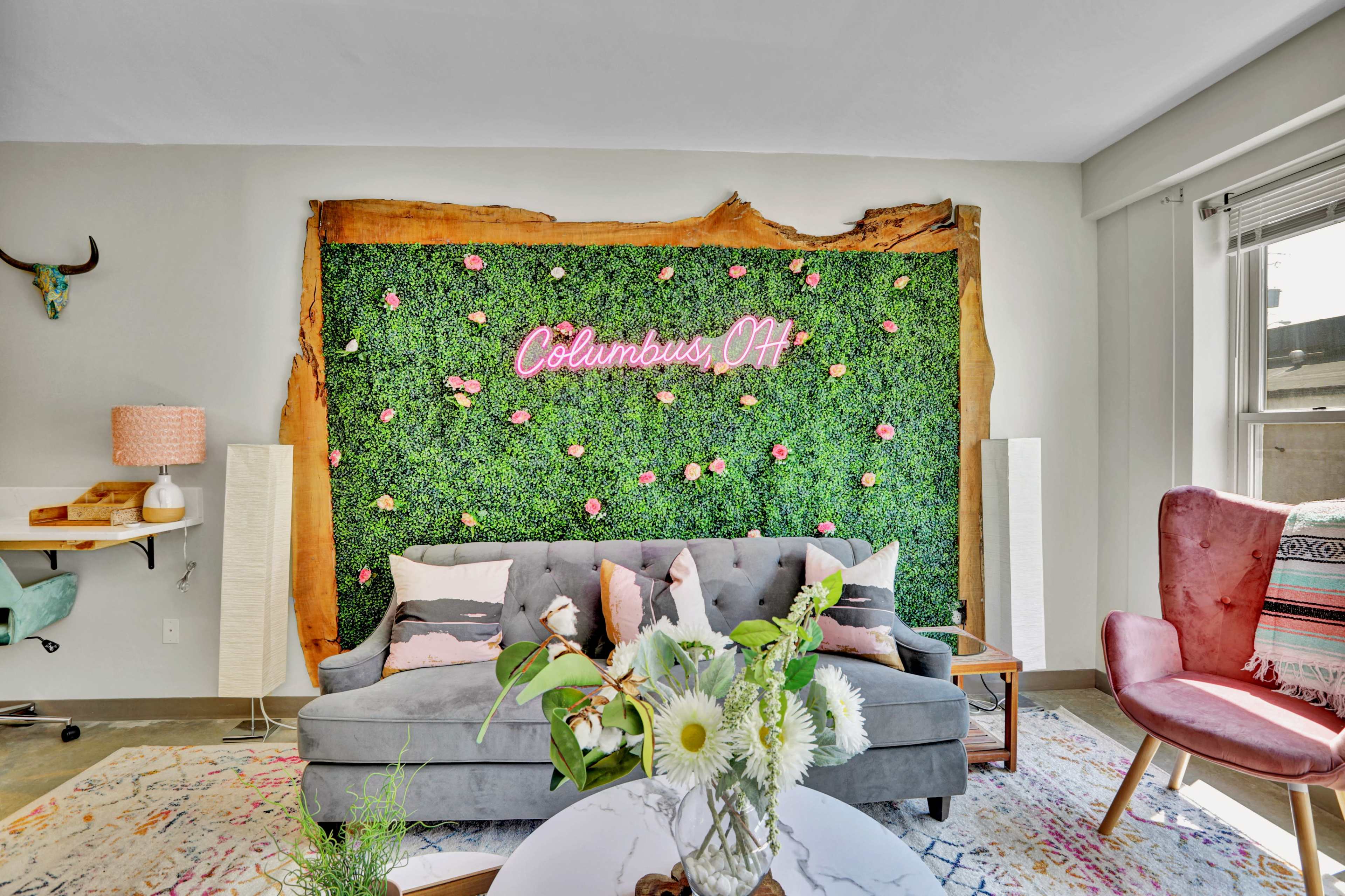 A living room with a gray sofa in front of a green wall adorned with pink flowers and a neon sign that reads "Columbus, OH."