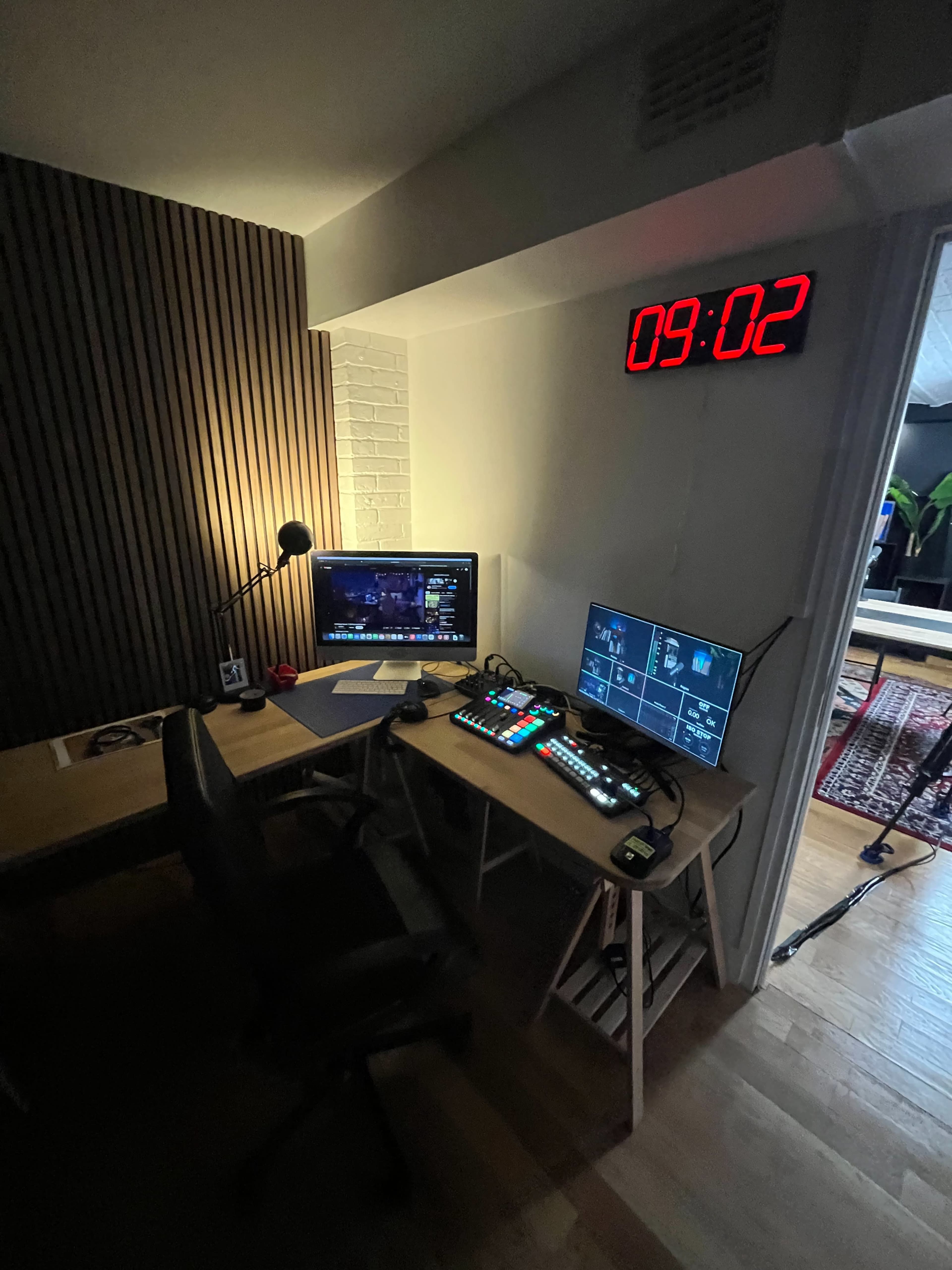 A modern workspace featuring a desk with a computer monitor, a second screen displaying controls, and a large digital clock on the wall.