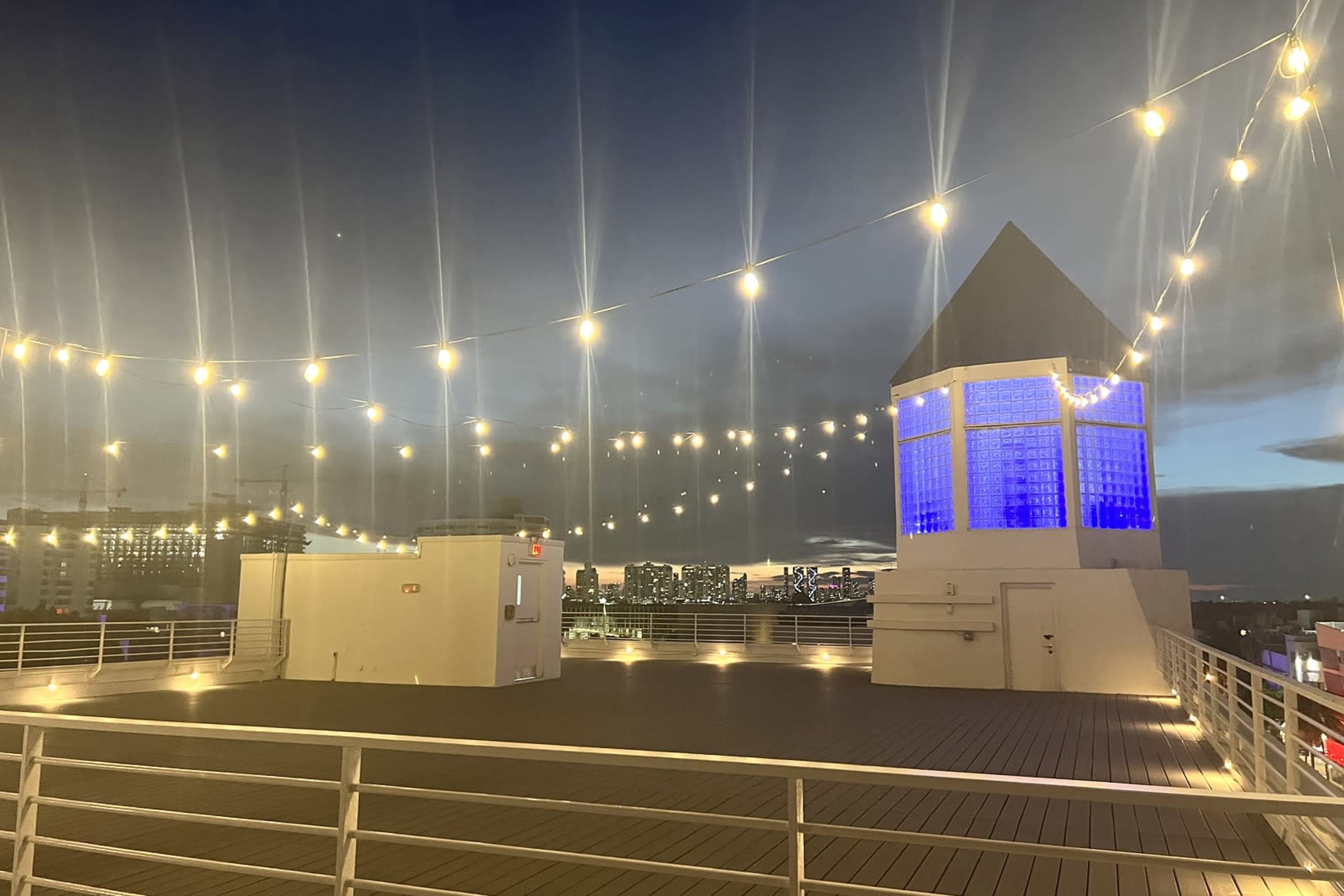 The image shows a rooftop area illuminated by string lights, with a blue-lit structure at one end and a city skyline in the background.