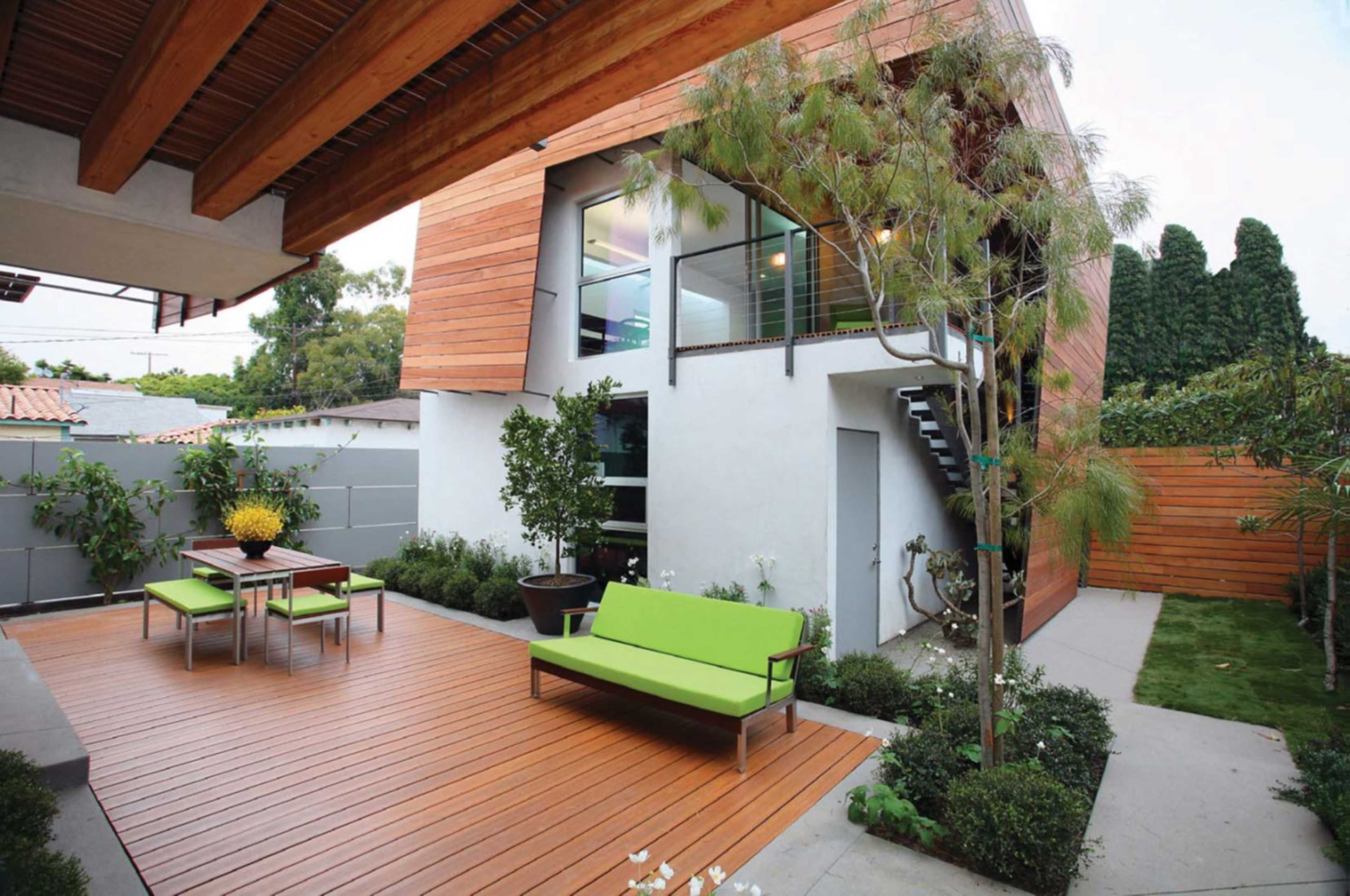 The image shows a modern backyard with a wooden deck, a green seating area, and a two-story building featuring large windows.