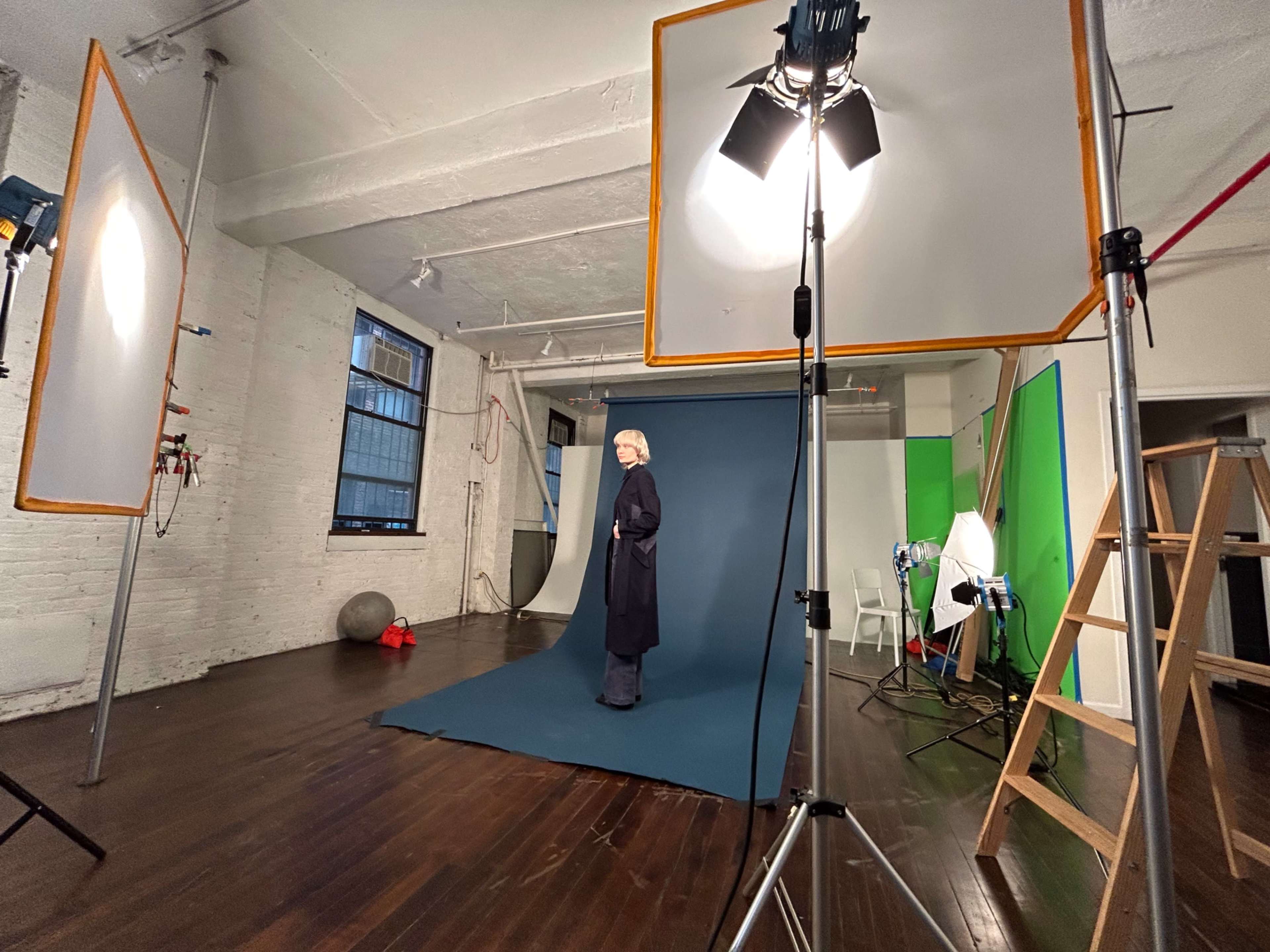 A model stands on a blue backdrop in a well-lit photography studio, surrounded by lighting equipment and props.