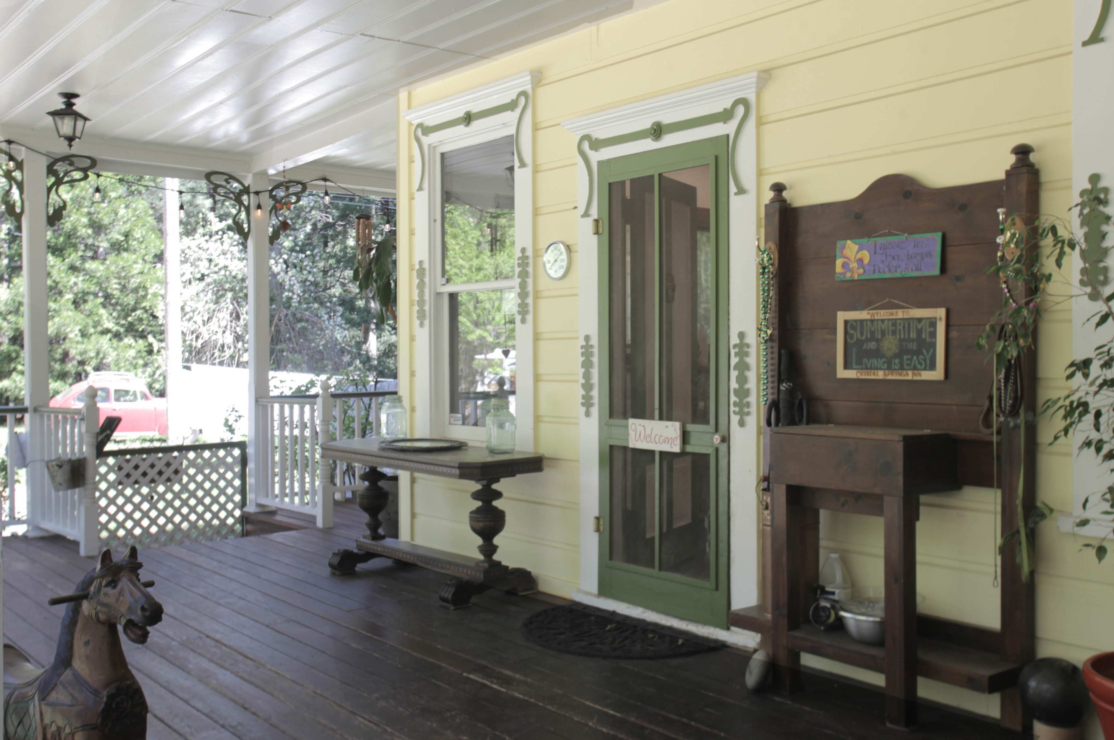 The image features a porch with yellow walls, a green door, a wooden table, and a cabinet displaying a sign.