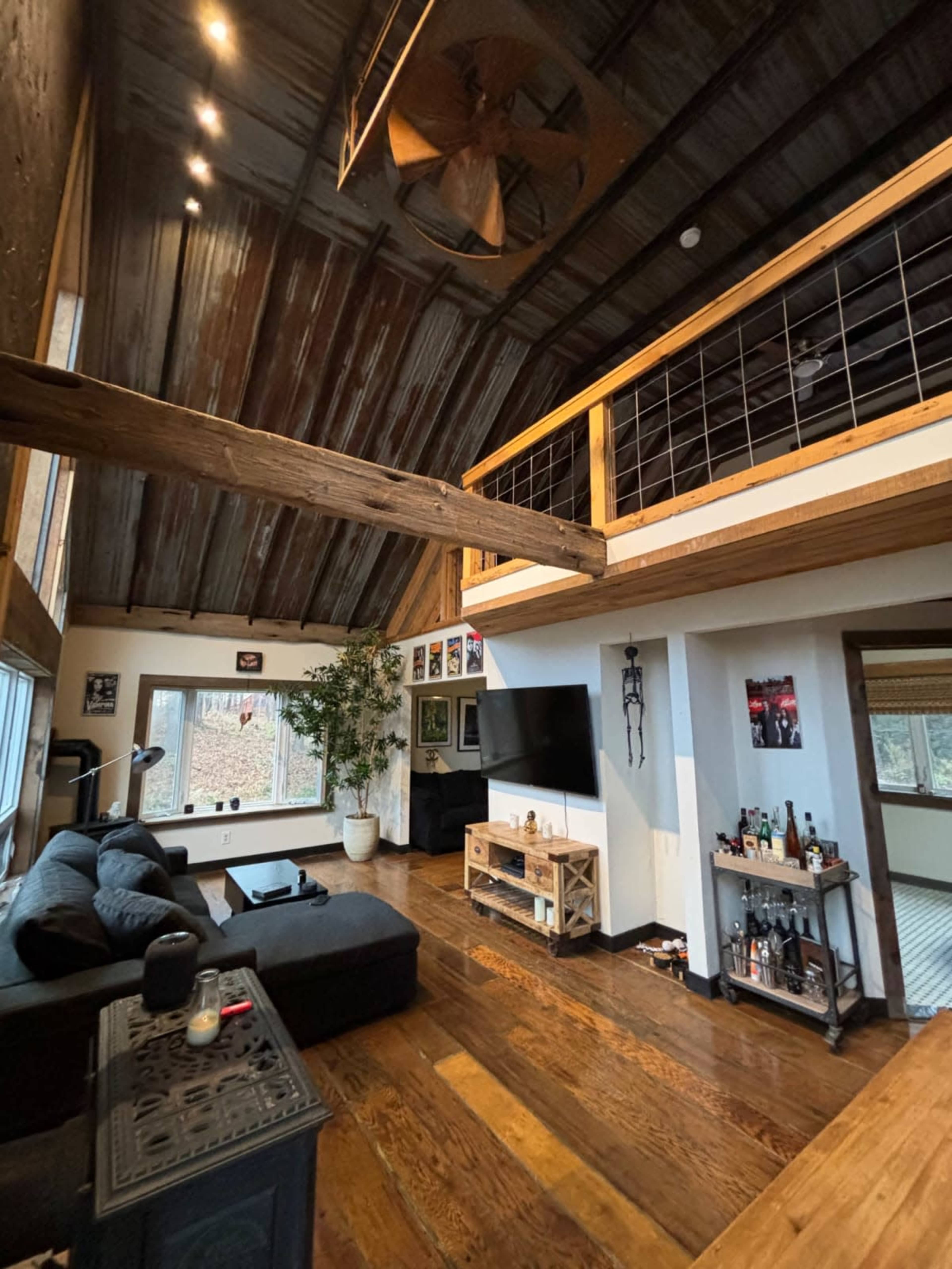 The image shows a spacious, modern living room with large windows, wooden beams, and a lofted area overlooking the space.