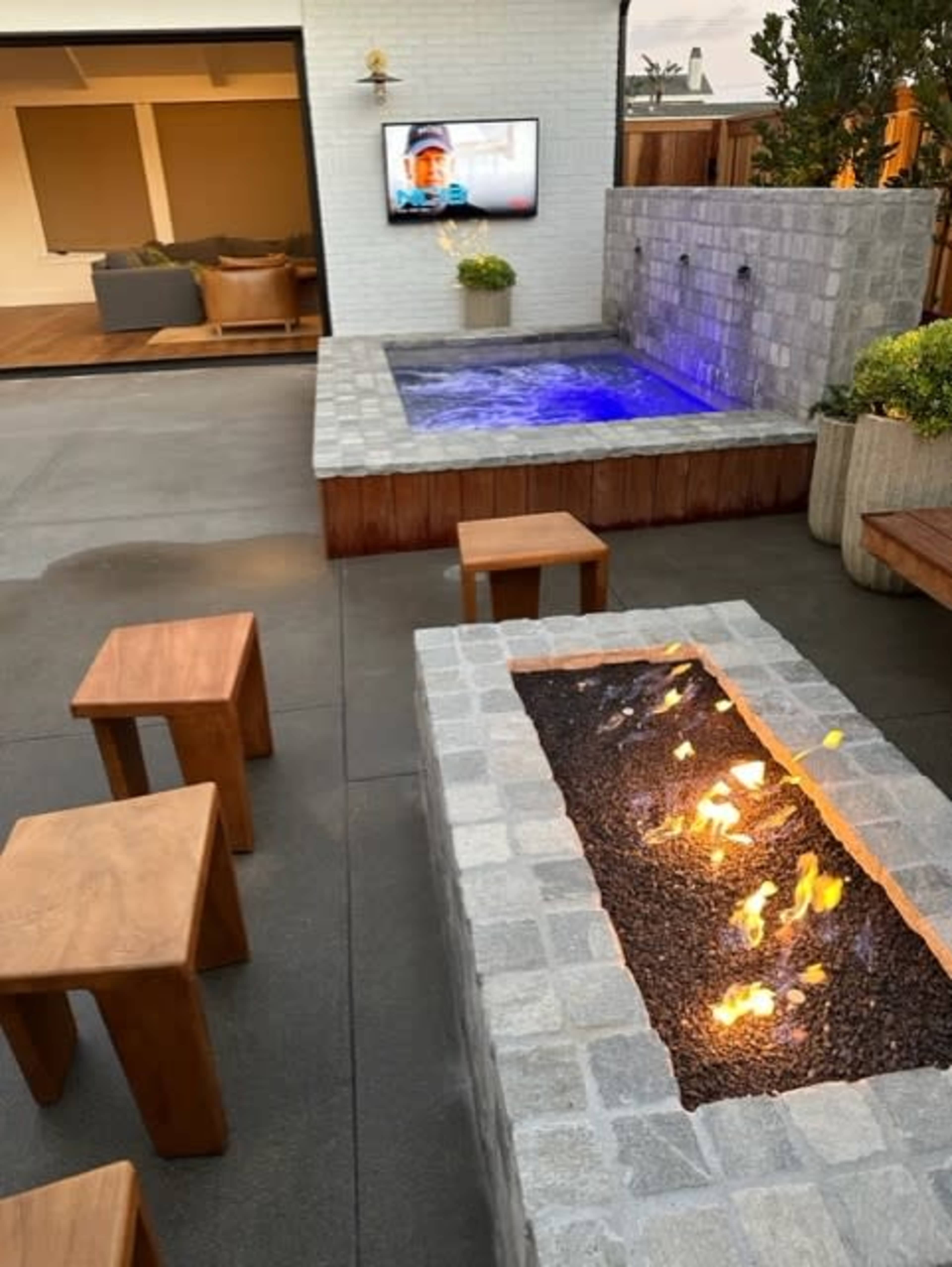 The image shows a modern outdoor patio with a hot tub, a fire pit, wooden seating, and a wall-mounted TV.