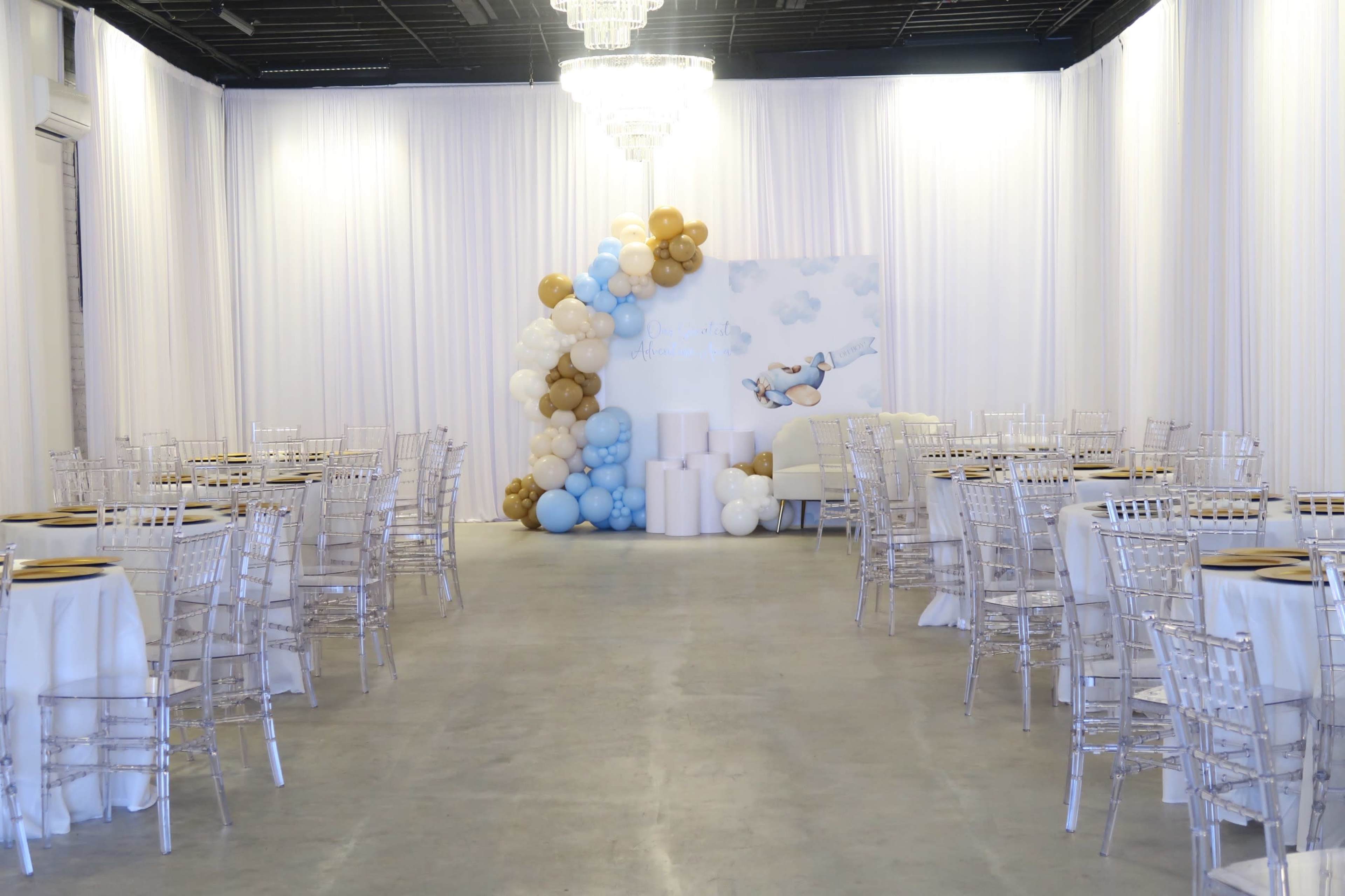 A spacious event venue features elegantly arranged round tables with white tablecloths and clear chiavari chairs, adorned with a backdrop of blue and gold balloons.