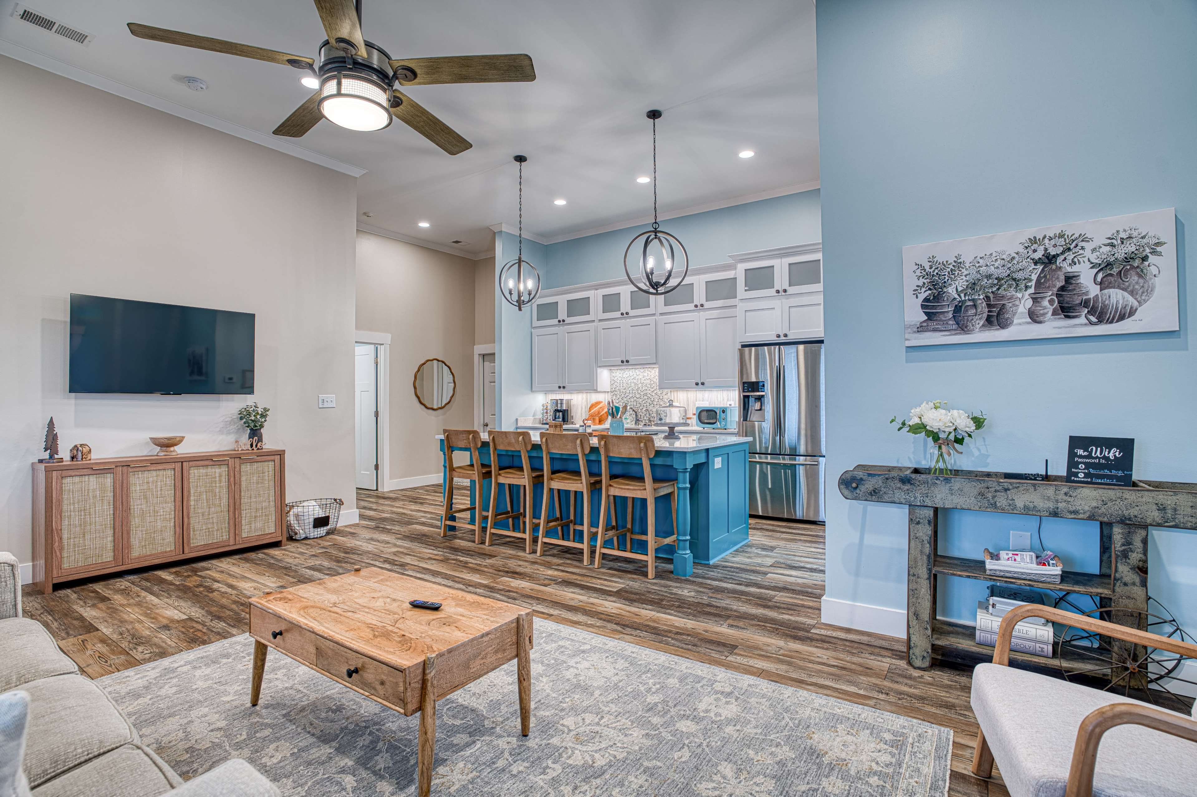 The image shows a modern living room and kitchen area featuring a light-colored sofa, a wooden coffee table, and a kitchen island with bar stools.