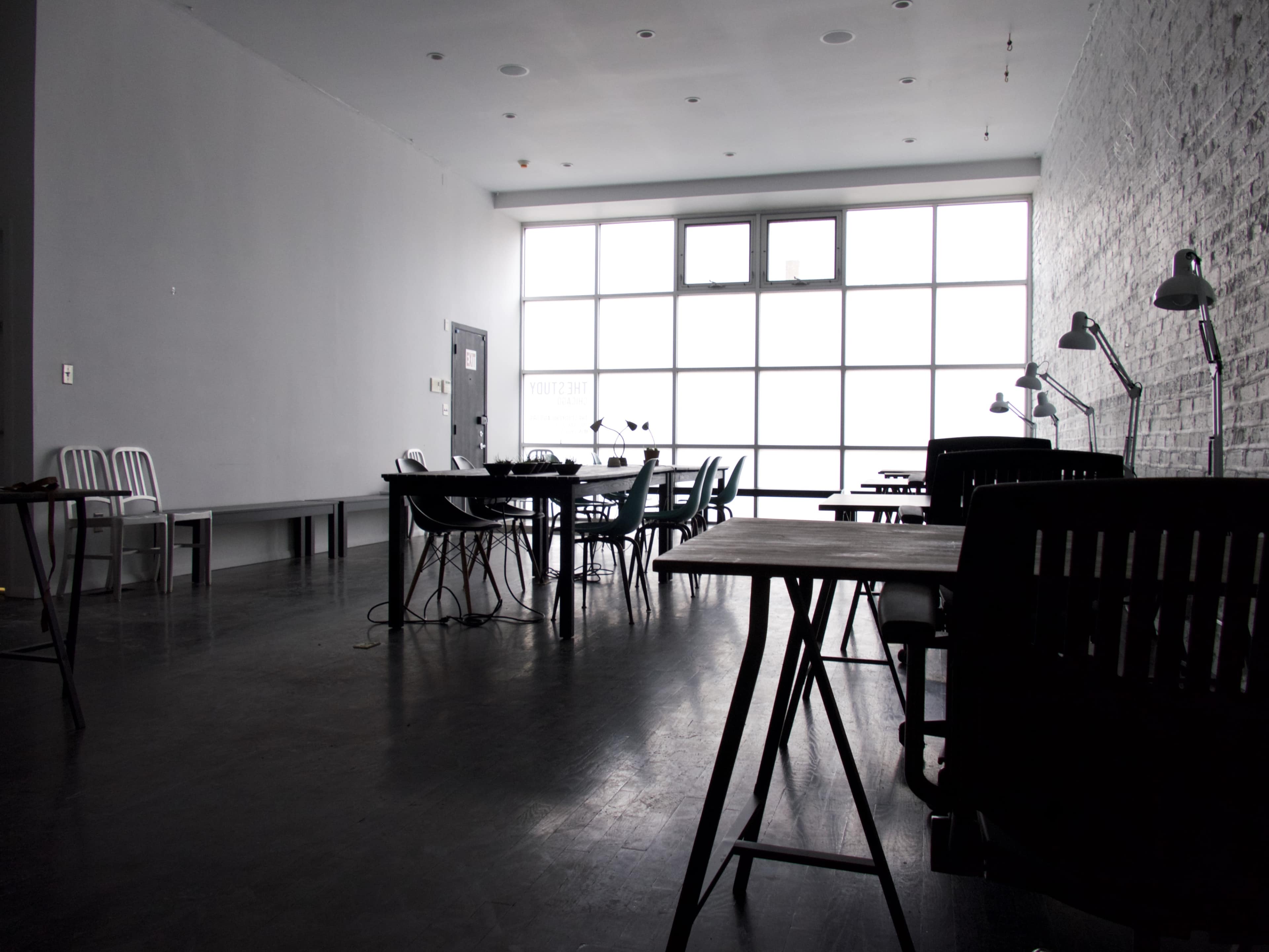 A spacious, modern room with multiple tables and chairs, featuring large windows that allow natural light to enter.