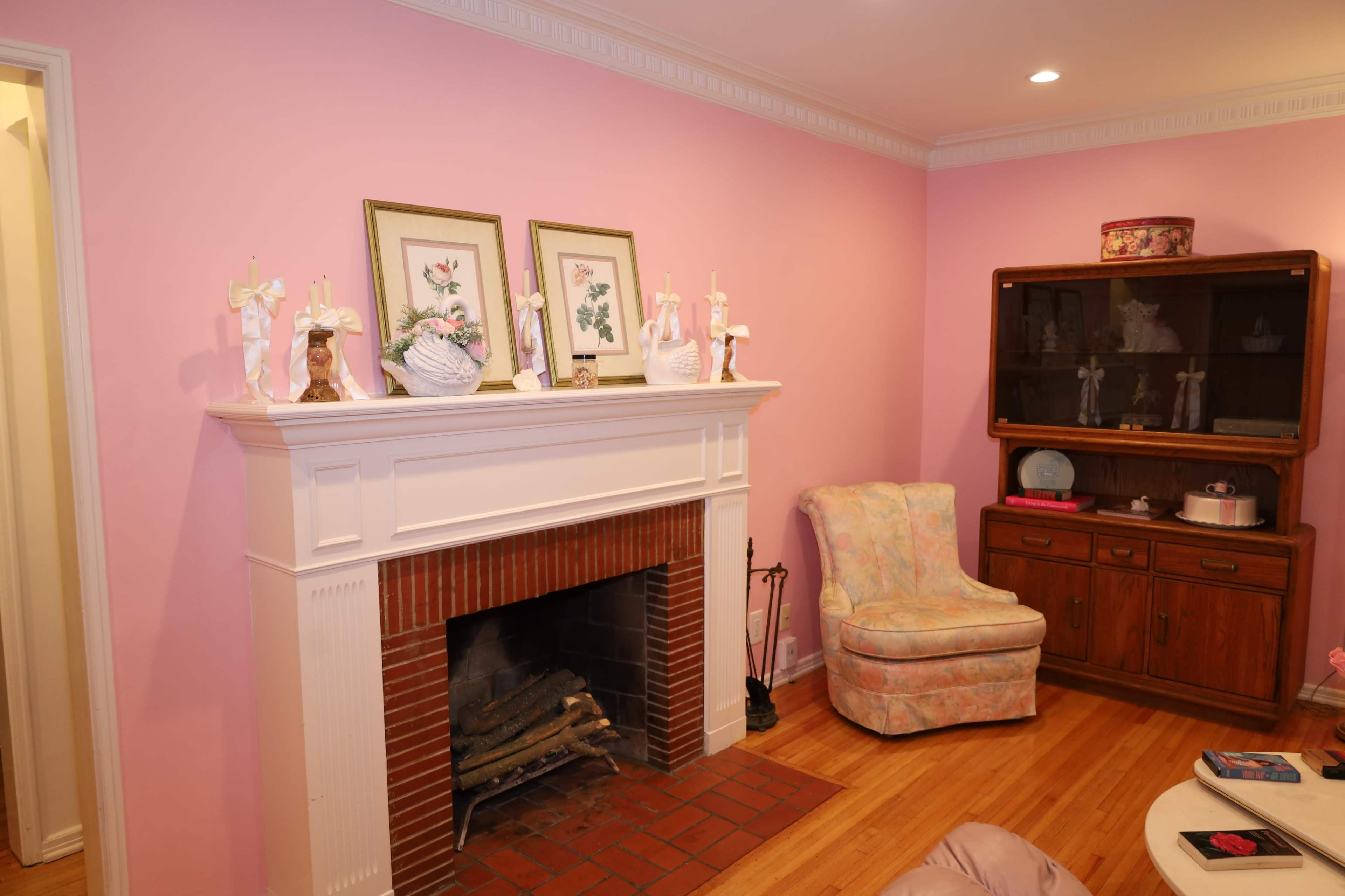 The image shows a cozy room with pink walls, a brick fireplace, a floral-decorated armchair, and wooden cabinetry.