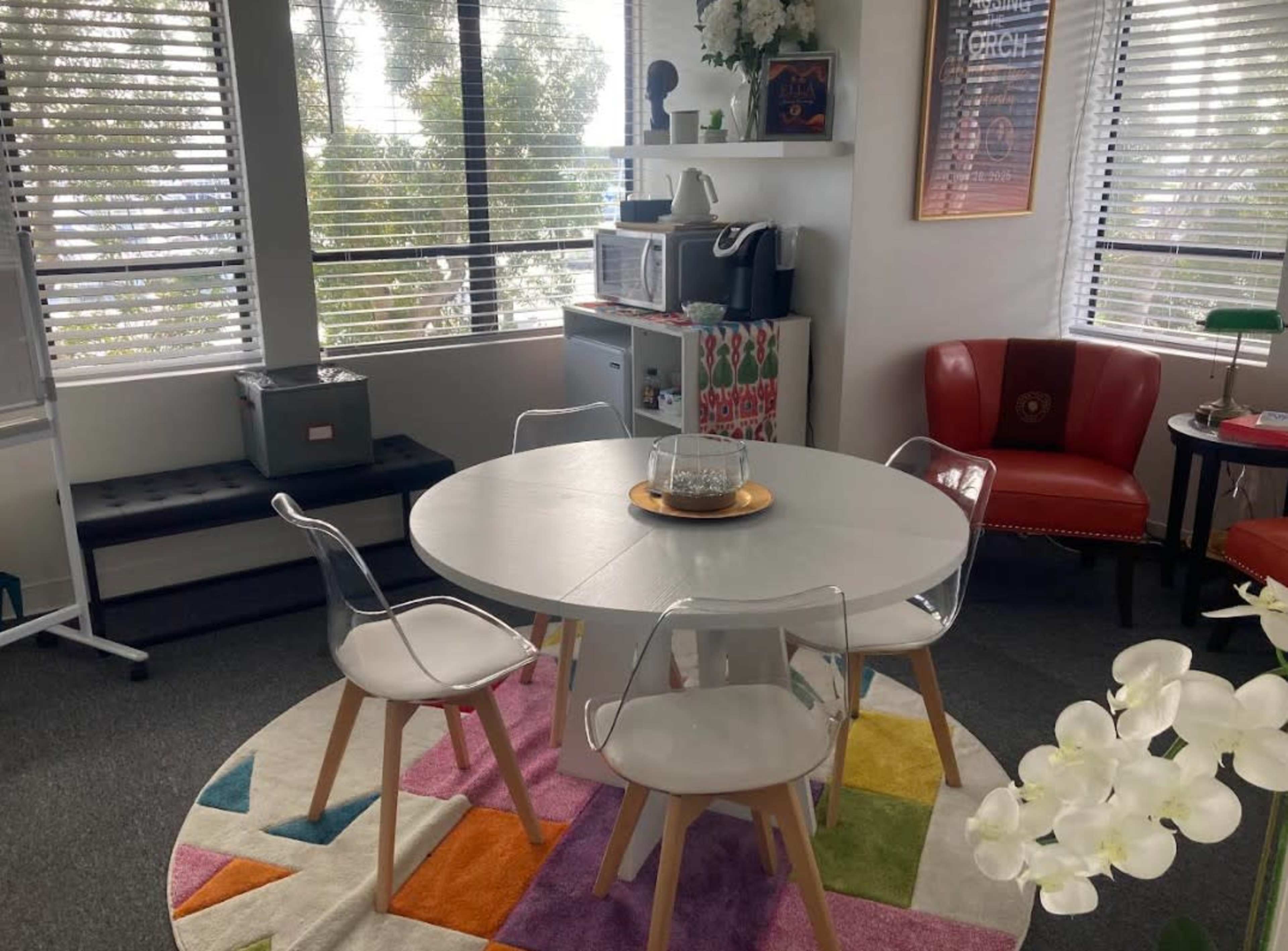 The image shows a modern office space featuring a round white table surrounded by four transparent chairs, with a colorful rug, a microwave on a shelf, and large windows providing natural light.