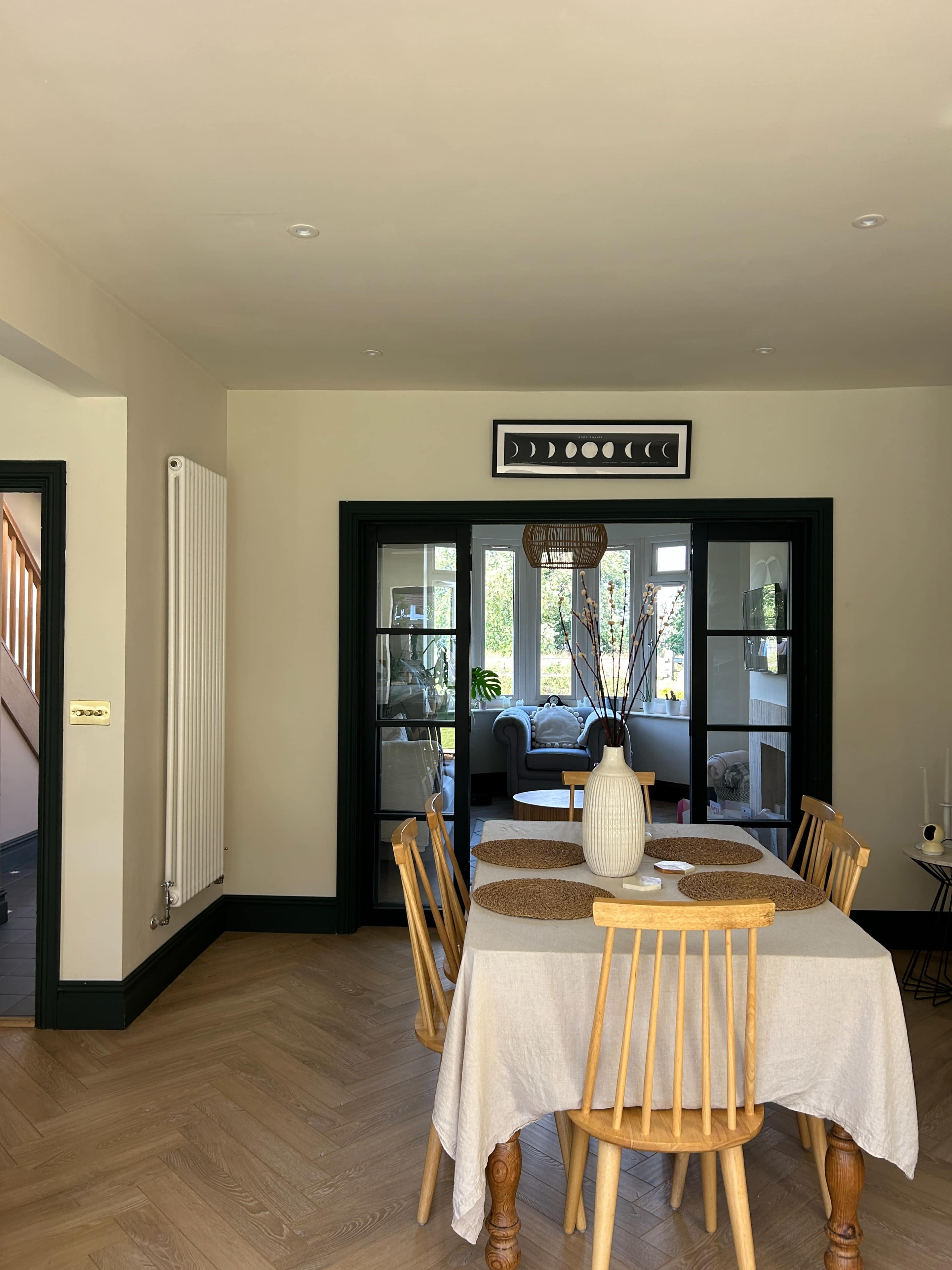 A dining area features a light wood table surrounded by six wooden chairs, with a decorative vase and placemats on the table, and a view of a living room through glass doors.