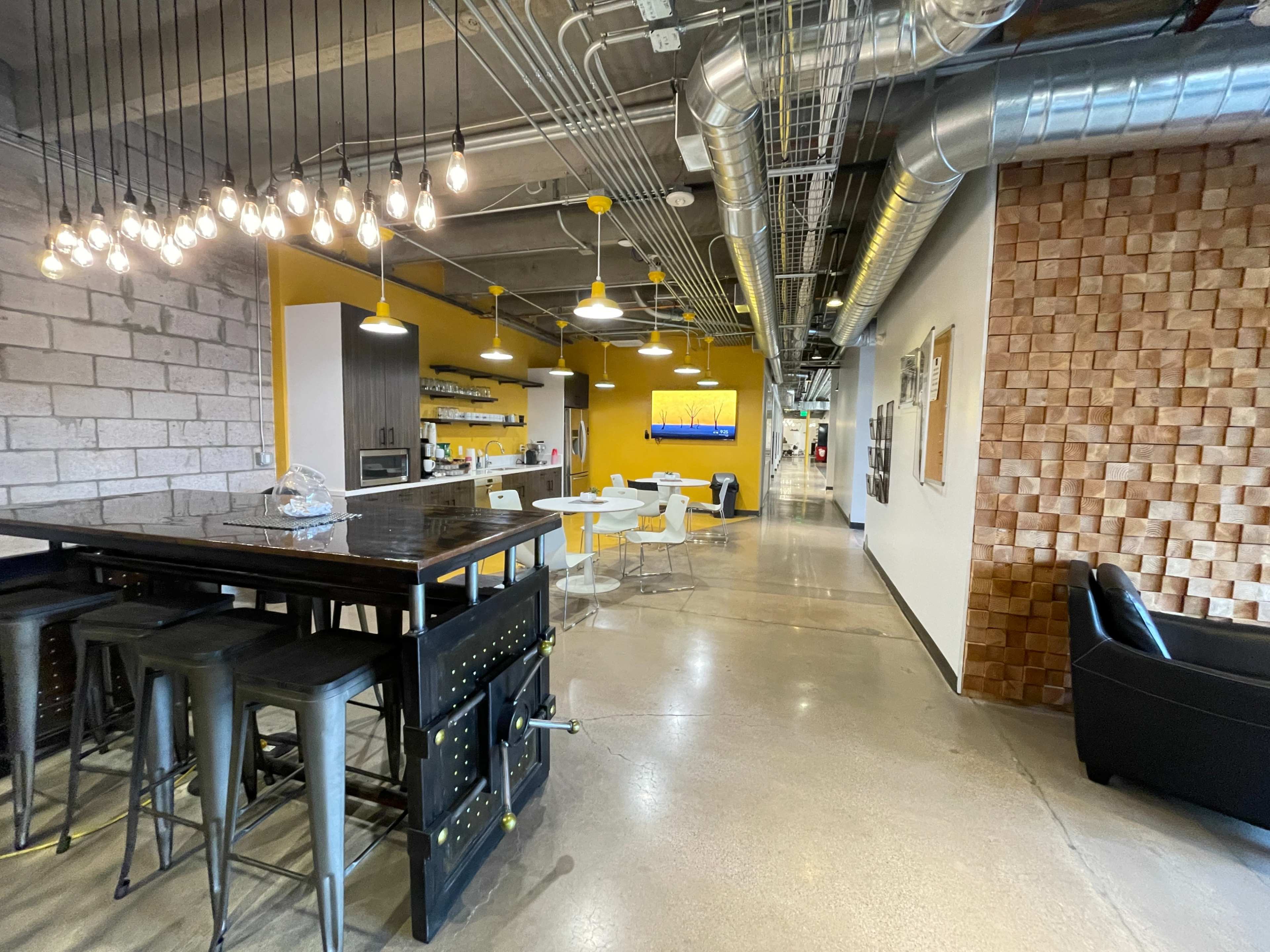 The image shows an open office space with a kitchen area featuring a large bar table and modern seating, alongside a hallway leading to additional workspaces.