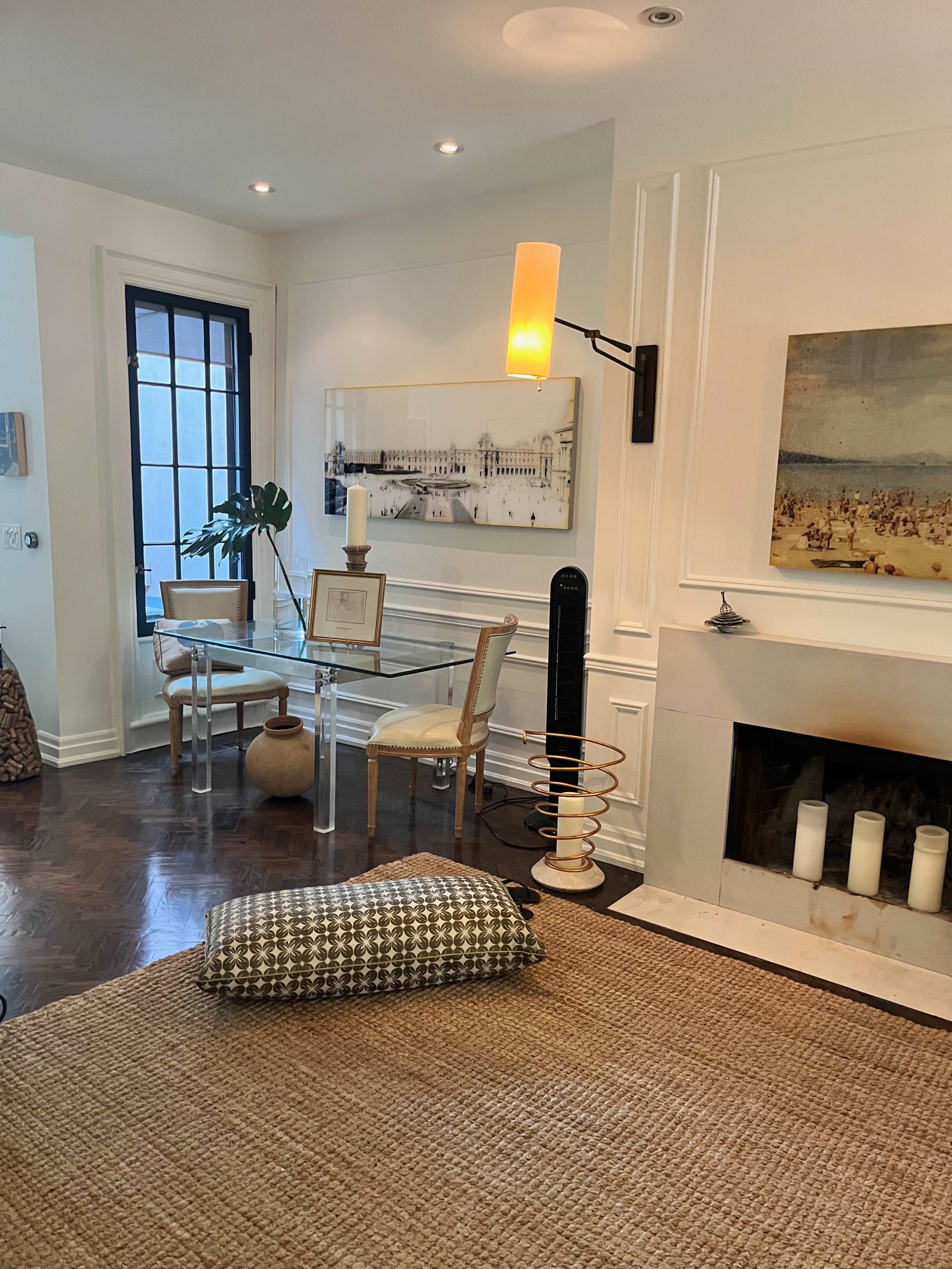 A modern living room with a glass dining table, a wall-mounted lamp, and a fireplace featuring several candles.