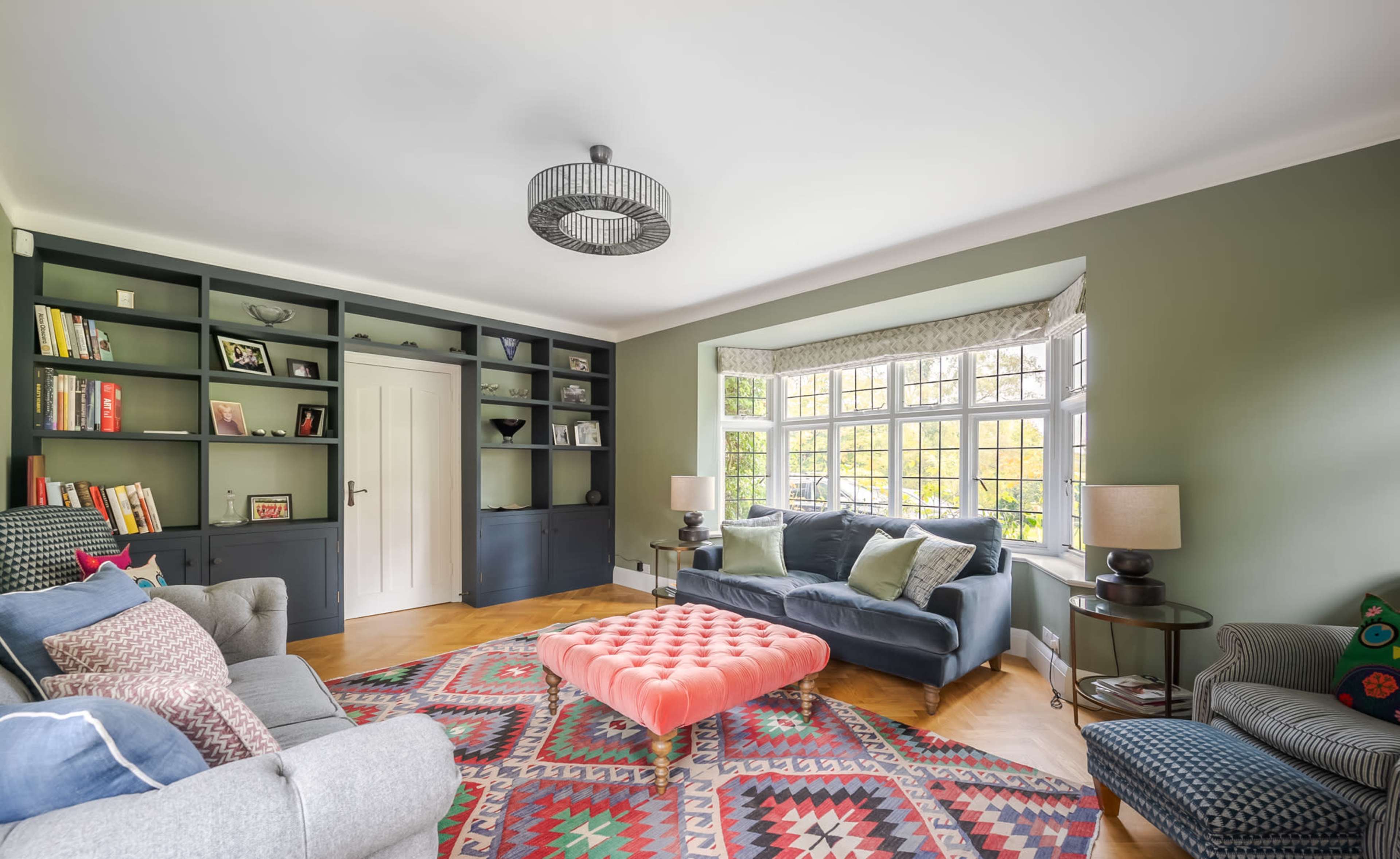 A cozy living room with a pink tufted coffee table, a green accent wall, and large windows letting in natural light.