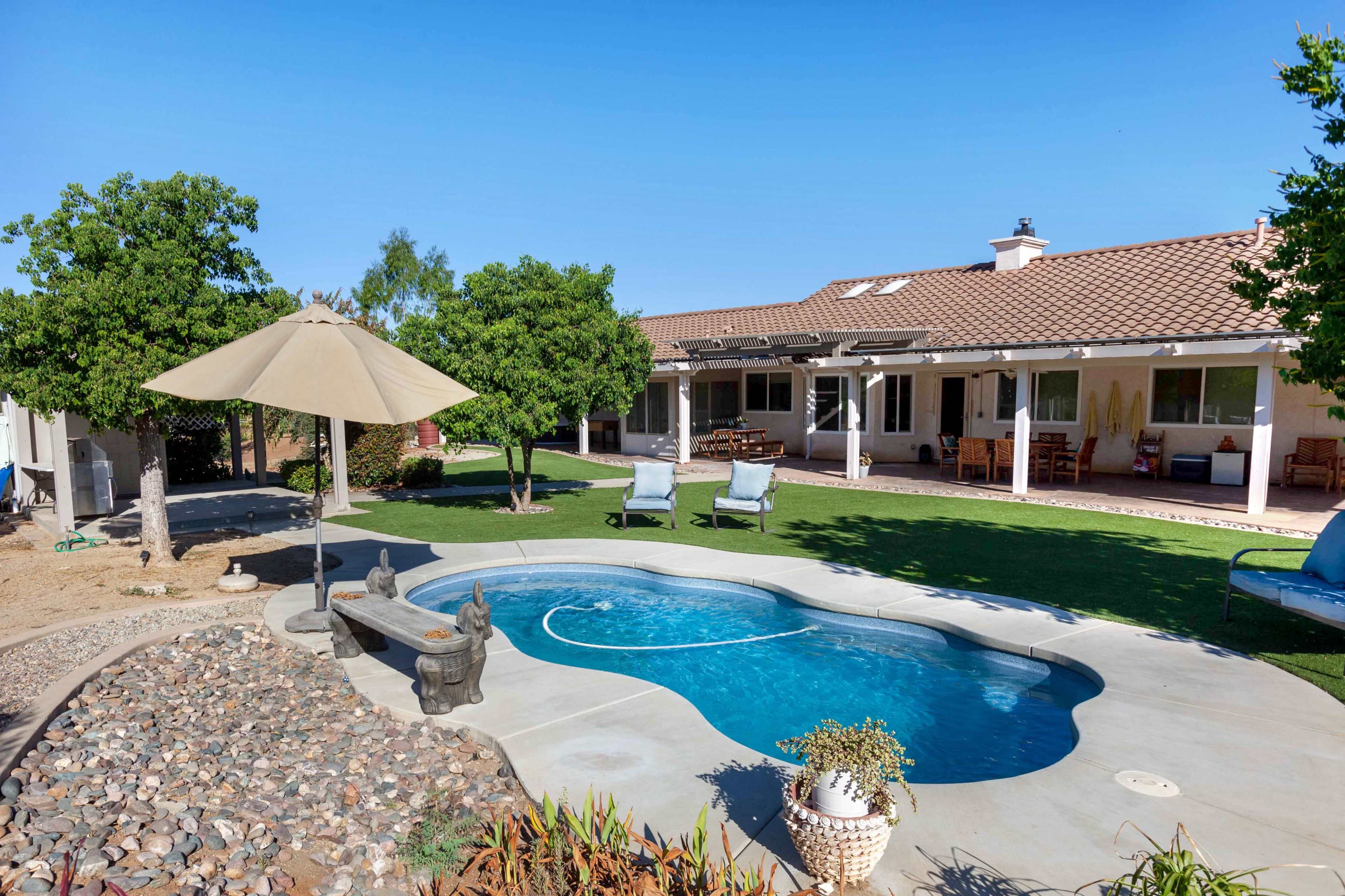 The image shows a landscaped backyard featuring a swimming pool, lounge chairs, and a patio area with shade provided by an umbrella.
