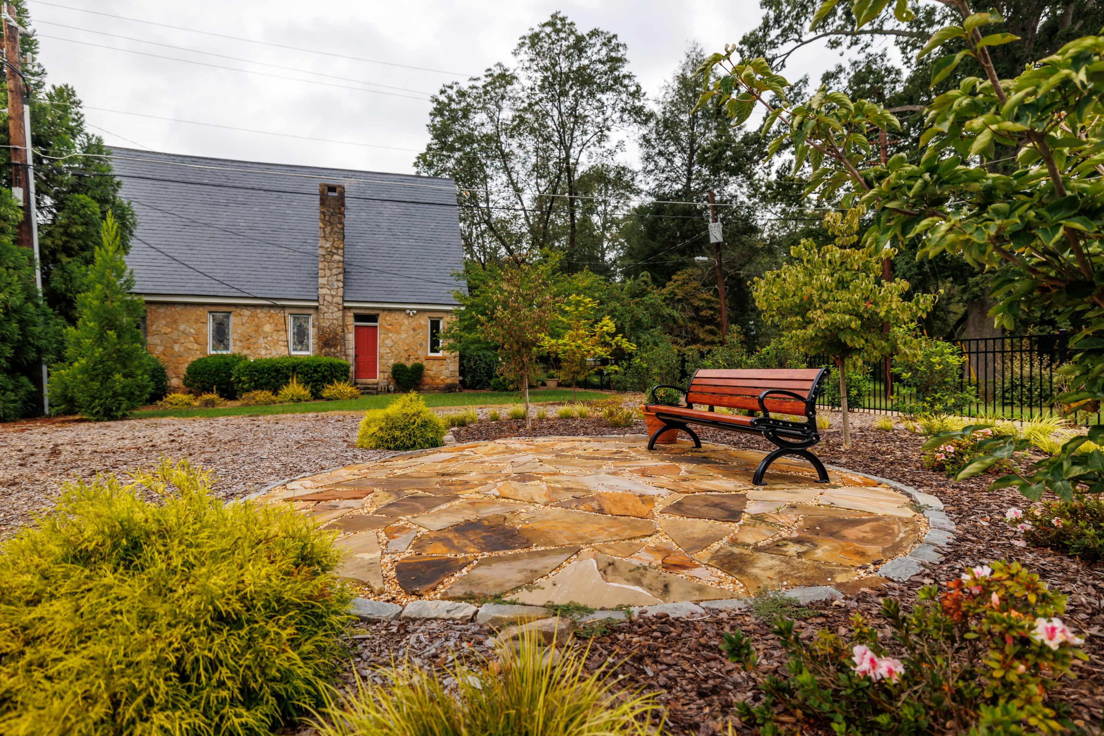 A stone patio with a bench is surrounded by landscaped plants and a house in the background.