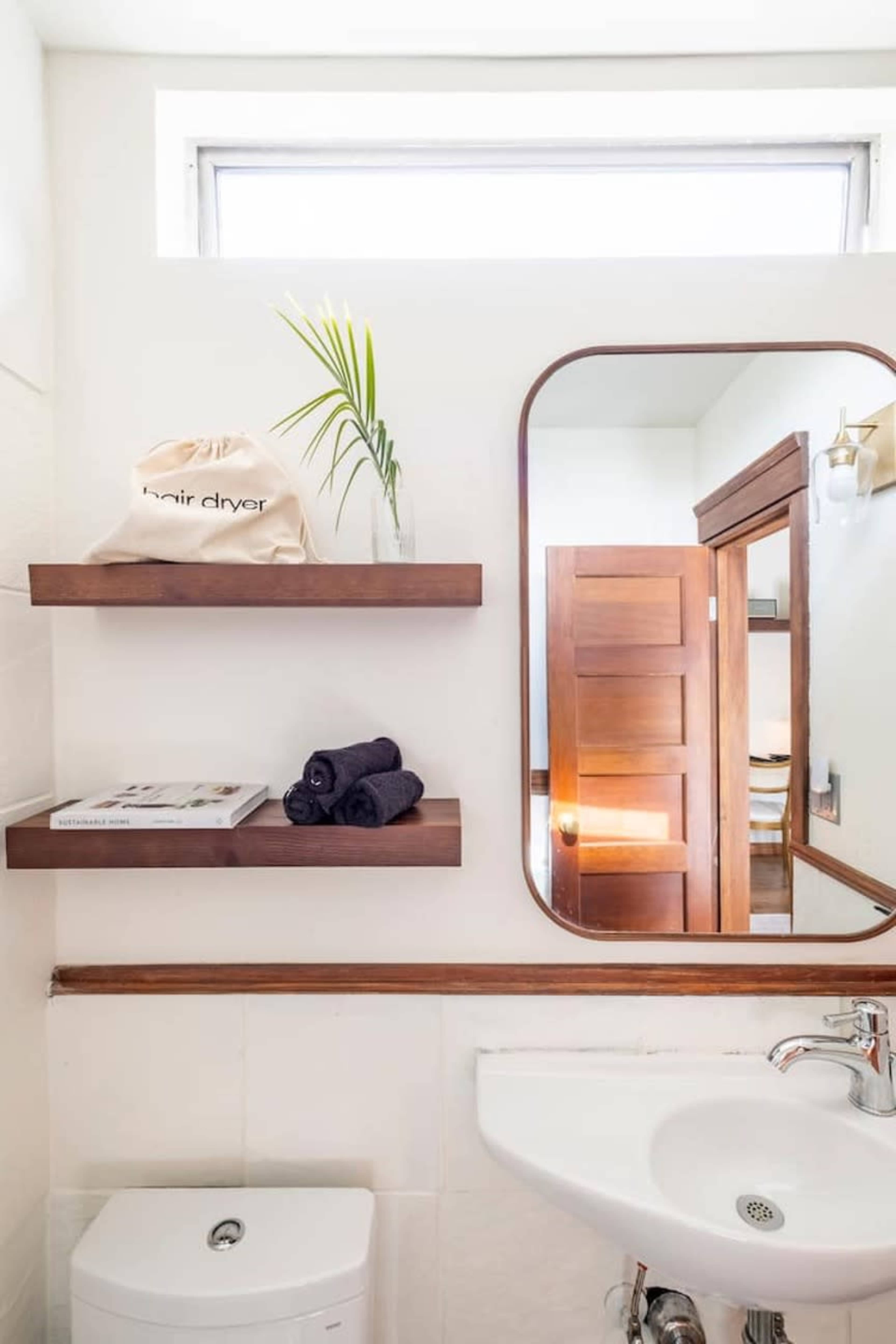 A bathroom with a sink, a round mirror, shelves holding towels and a hairdryer bag, and a wooden door slightly open in the background.