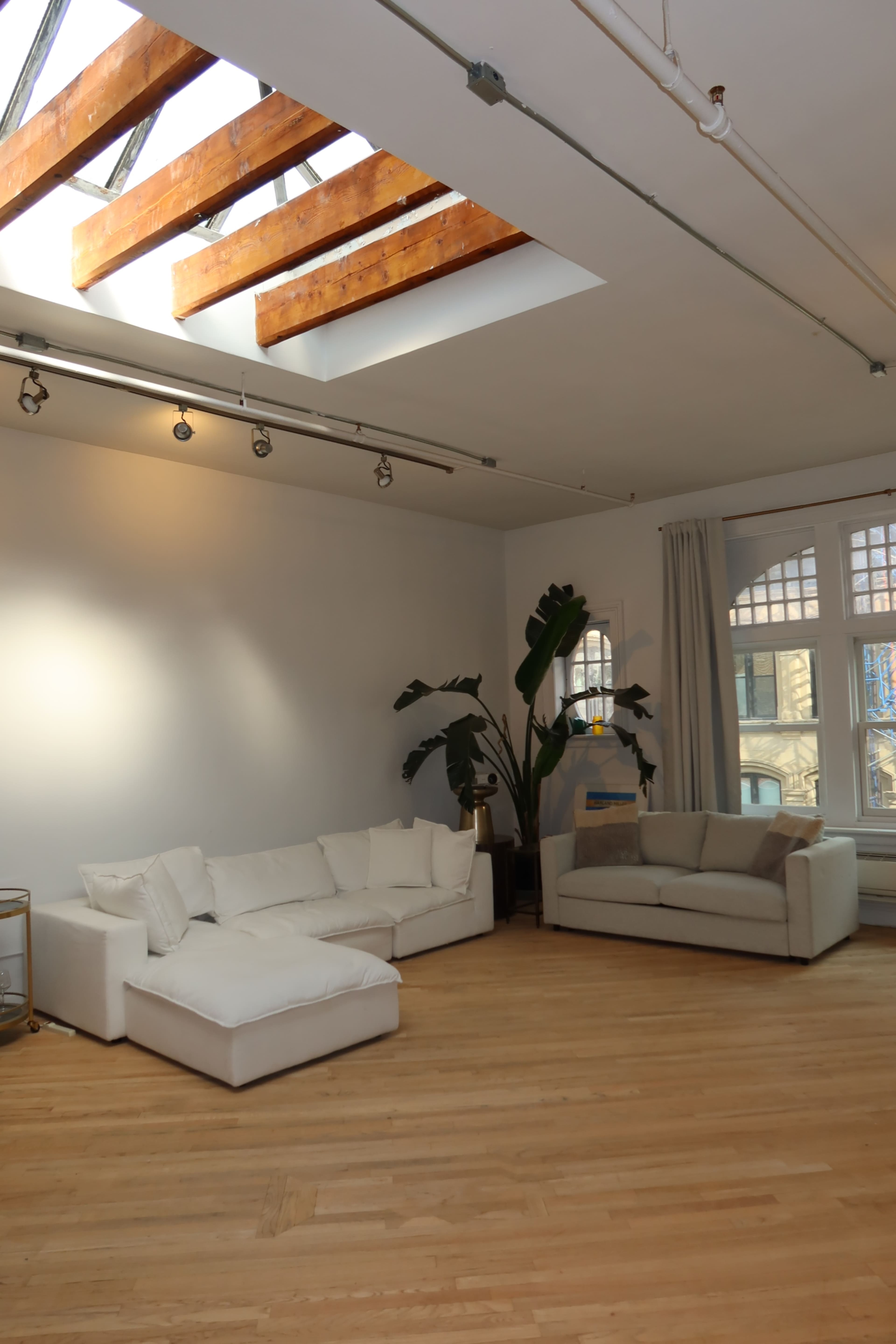 The image shows a spacious, bright living area with a white sectional sofa, a gray sofa, a large window with wooden beams, and potted plants.
