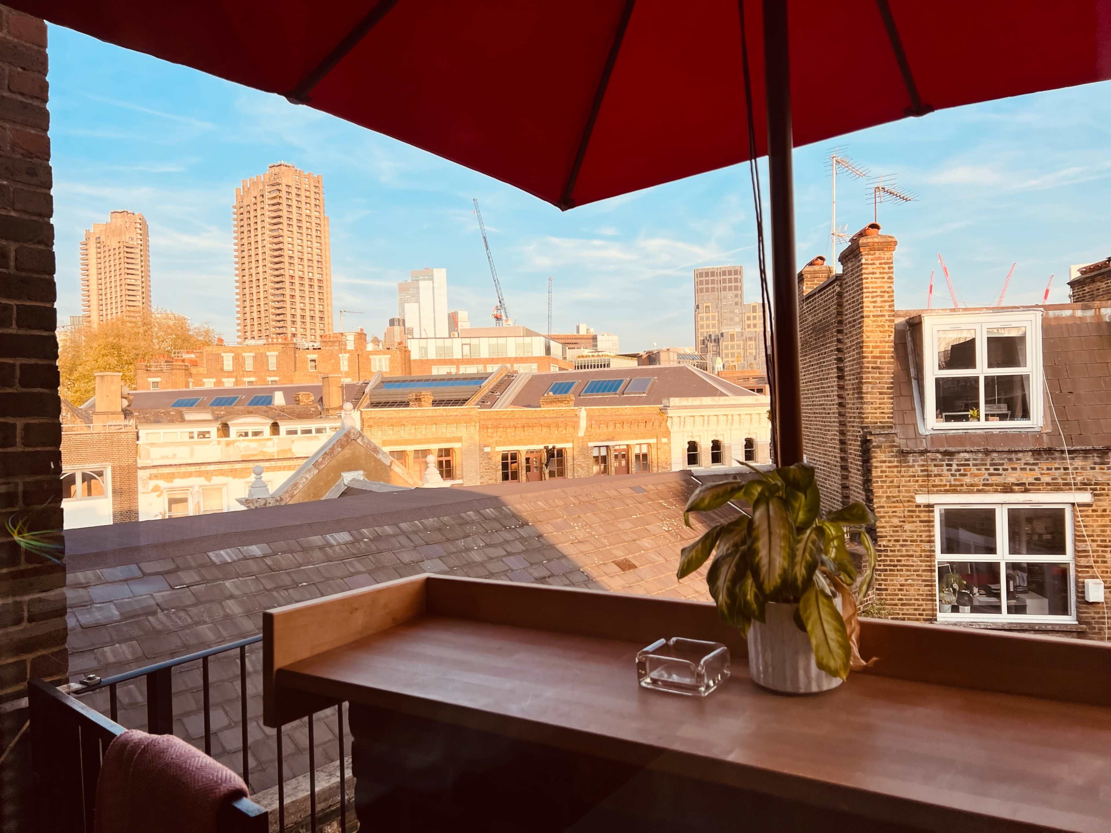 A wooden table with a small plant and an ashtray, overlooking rooftops and buildings under a red umbrella against a clear blue sky.