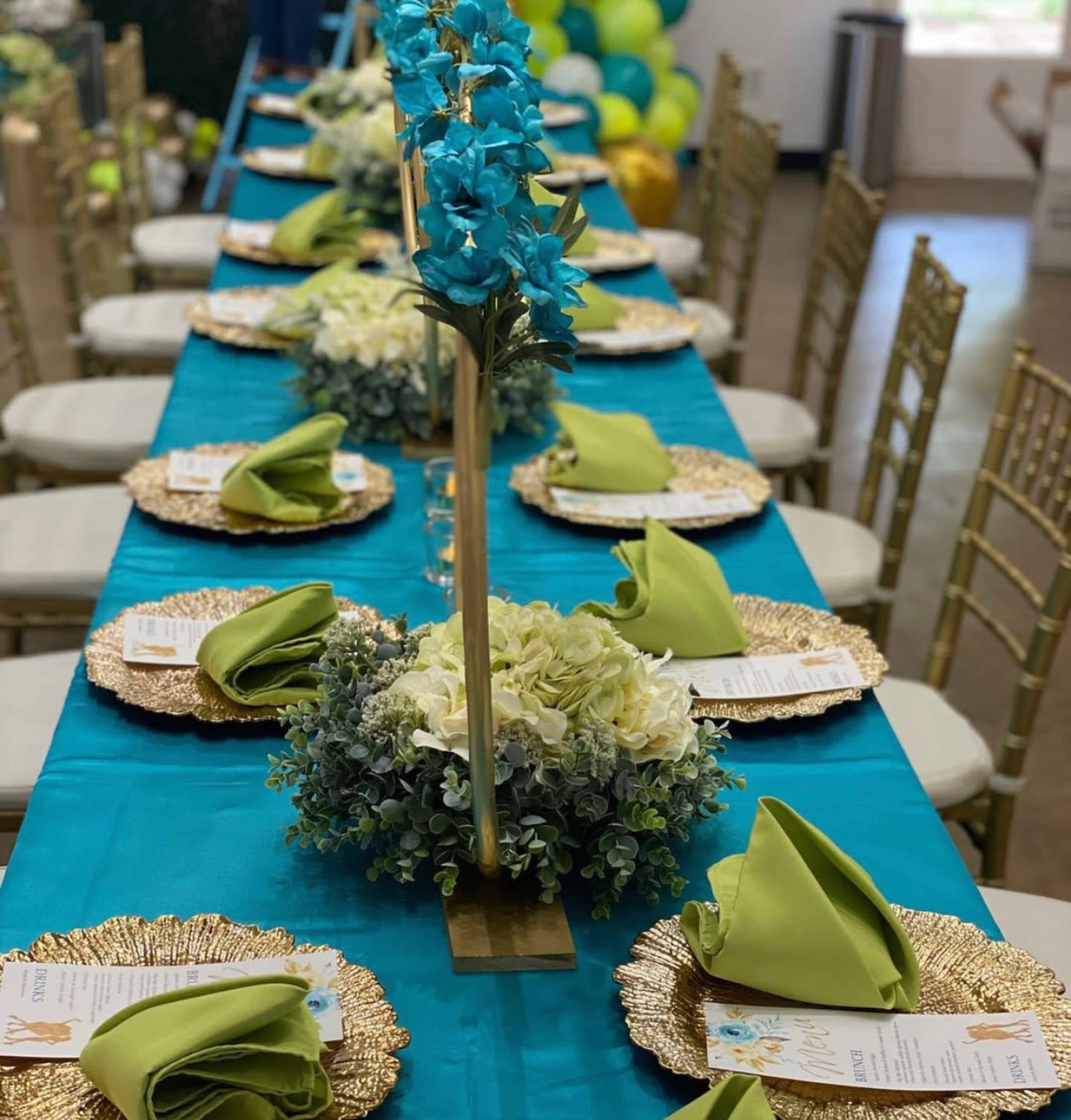 A long table set for a formal event features gold chargers, green napkins, and a floral centerpiece with blue orchids and white blooms.