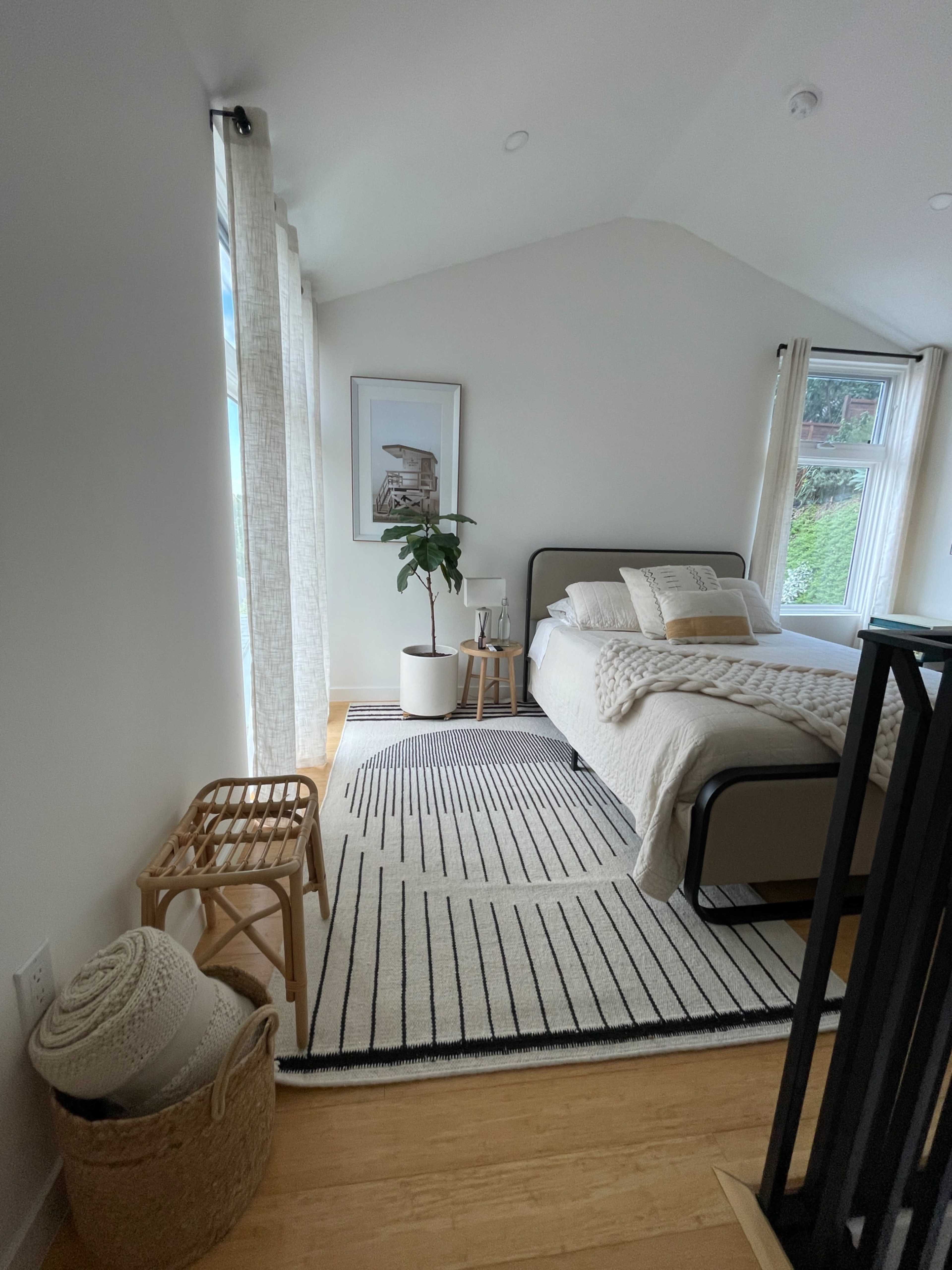 The image shows a well-lit bedroom featuring a bed with a patterned throw, a potted plant, a chair, and a rug on hardwood flooring.
