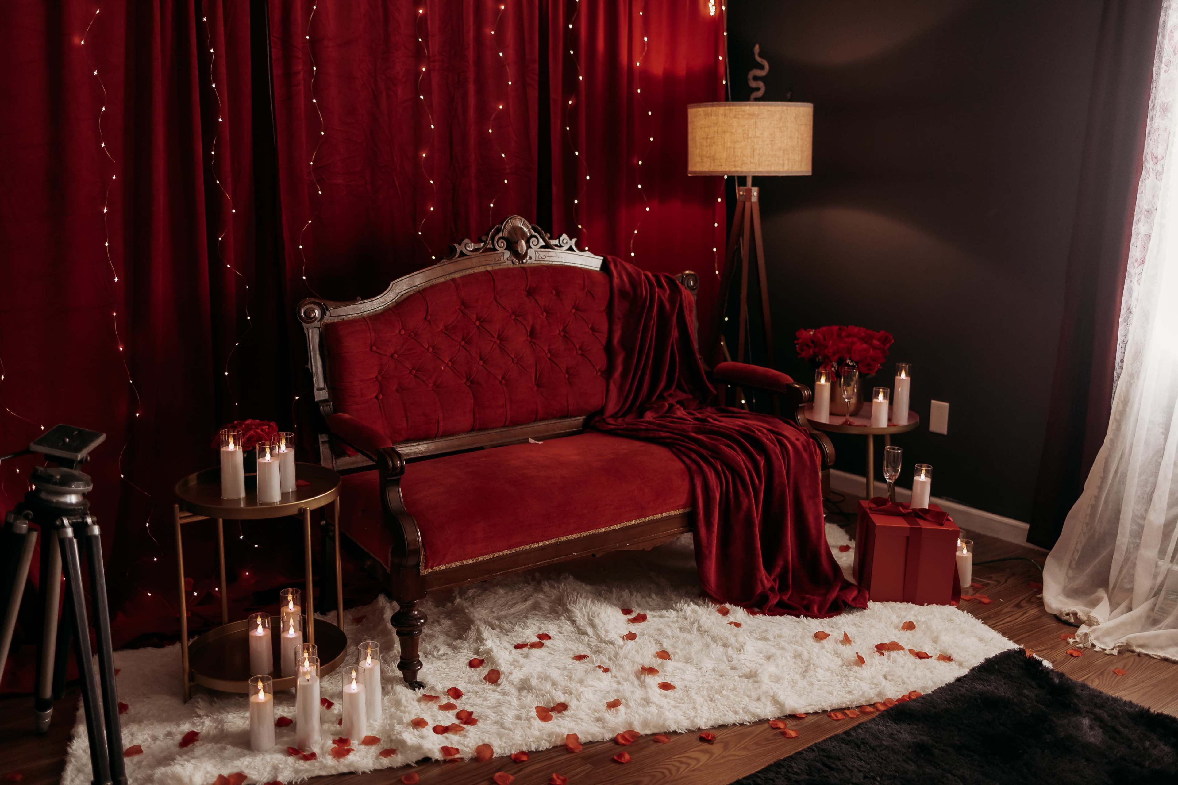 The scene features a richly decorated sitting area with a red velvet sofa, surrounded by candles, rose petals, and soft lighting against dark walls.