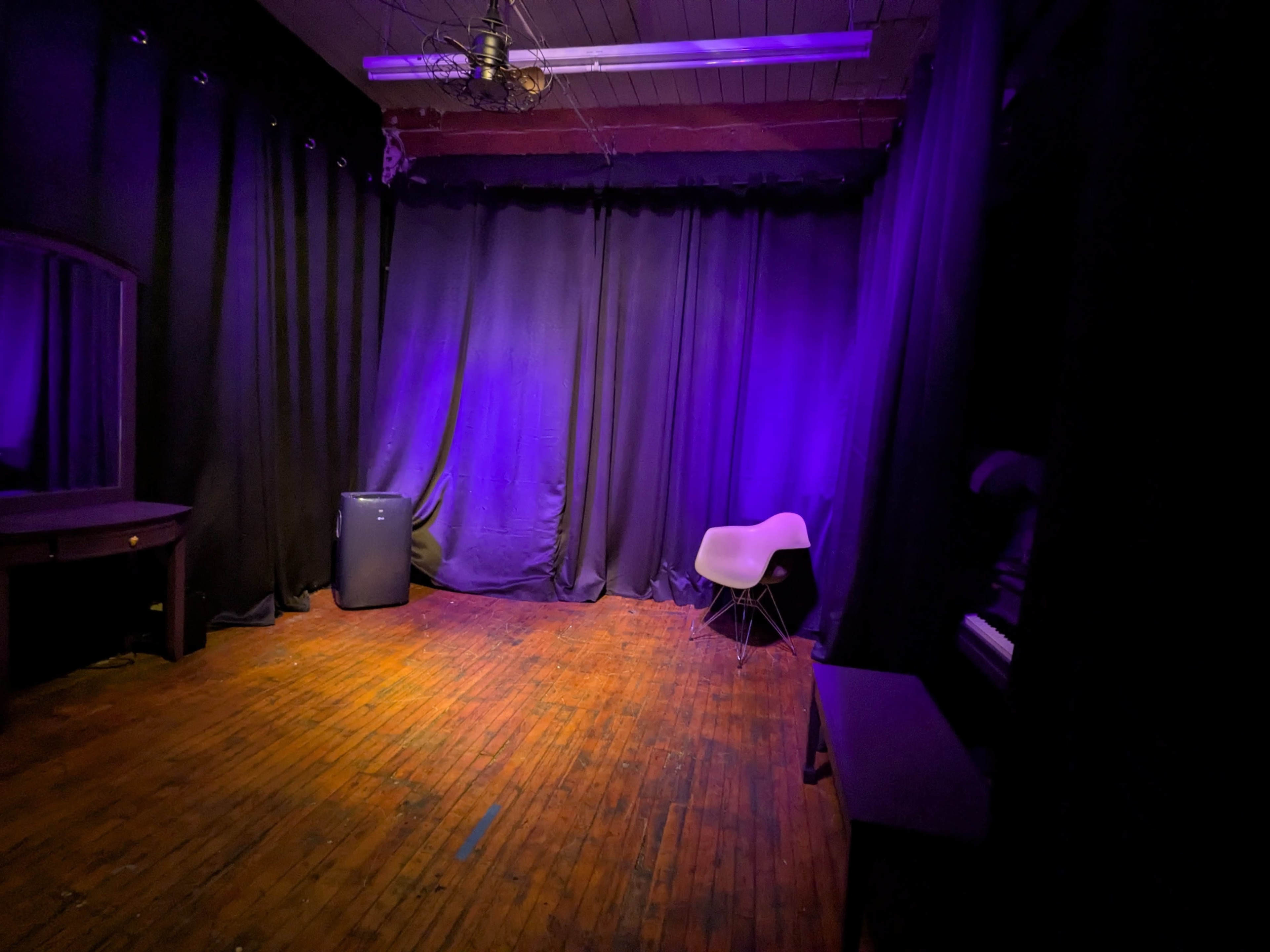 The image shows a dimly lit room with purple lighting, characterized by a wooden floor, a black curtain backdrop, and minimal furniture including a white chair and a small blue appliance.
