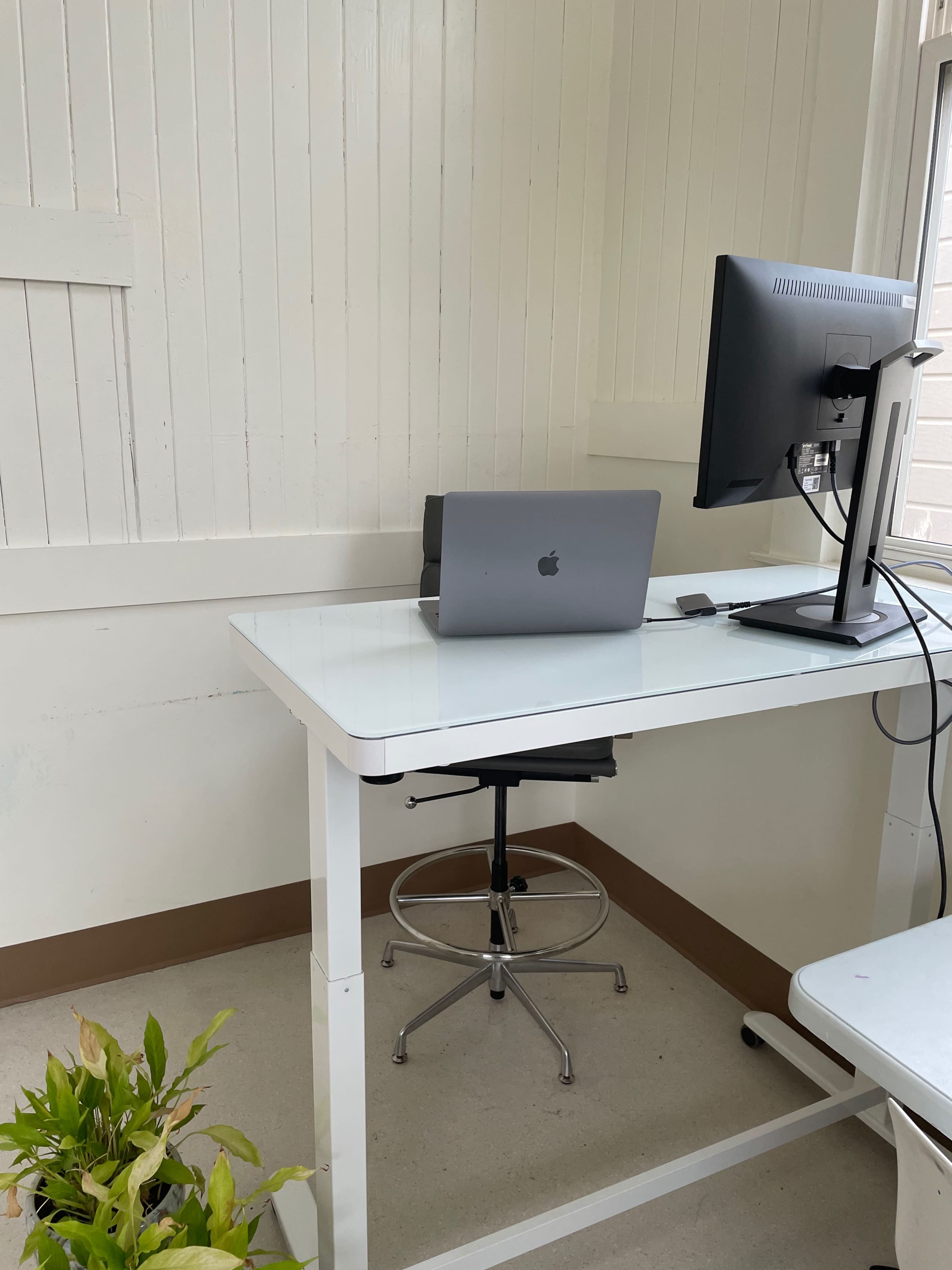 A modern workspace with a computer monitor and a laptop placed on a white desk, accompanied by a chair and a potted plant.