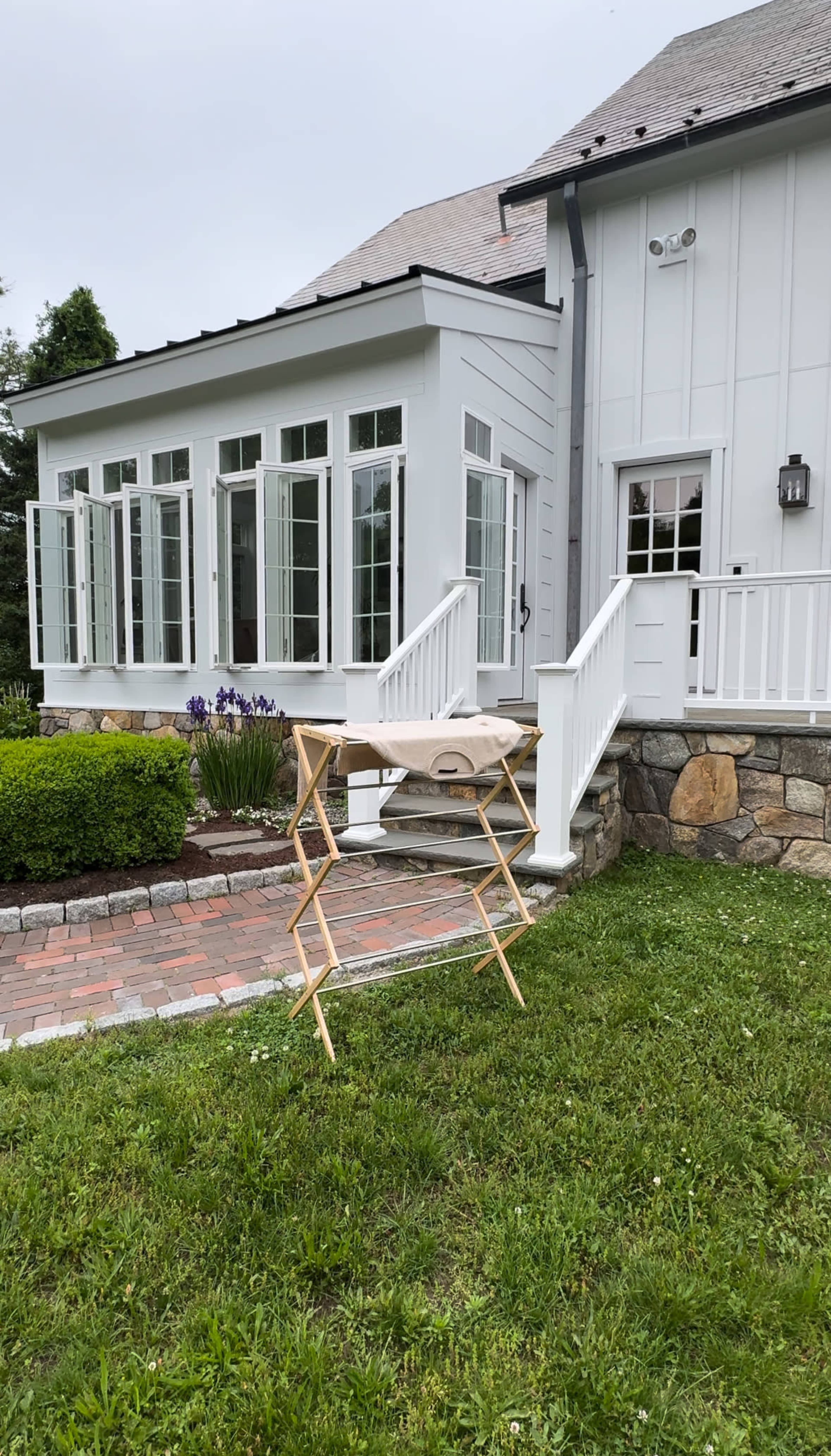 A wooden folding table is set up on the grass in front of a house with large windows and a brick pathway.