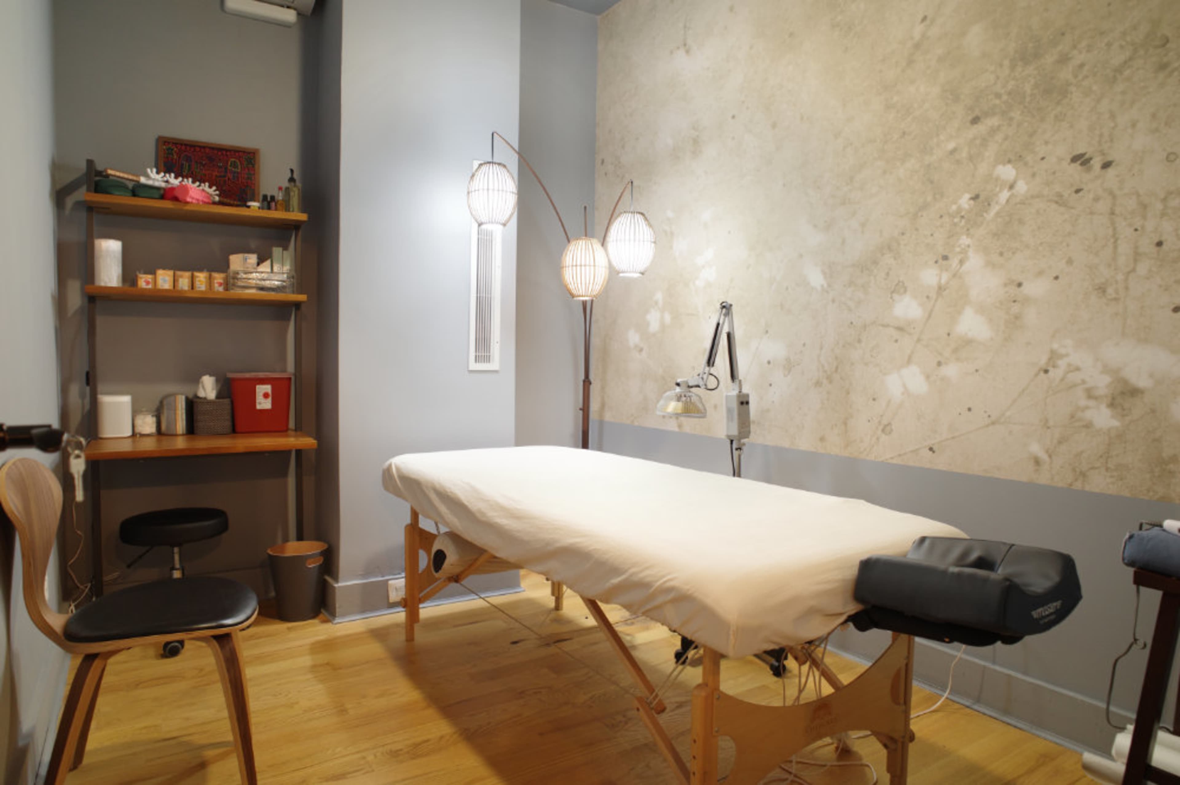 A massage room with a treatment table, a wooden shelf containing various items, and three floor lamps against a textured wall.