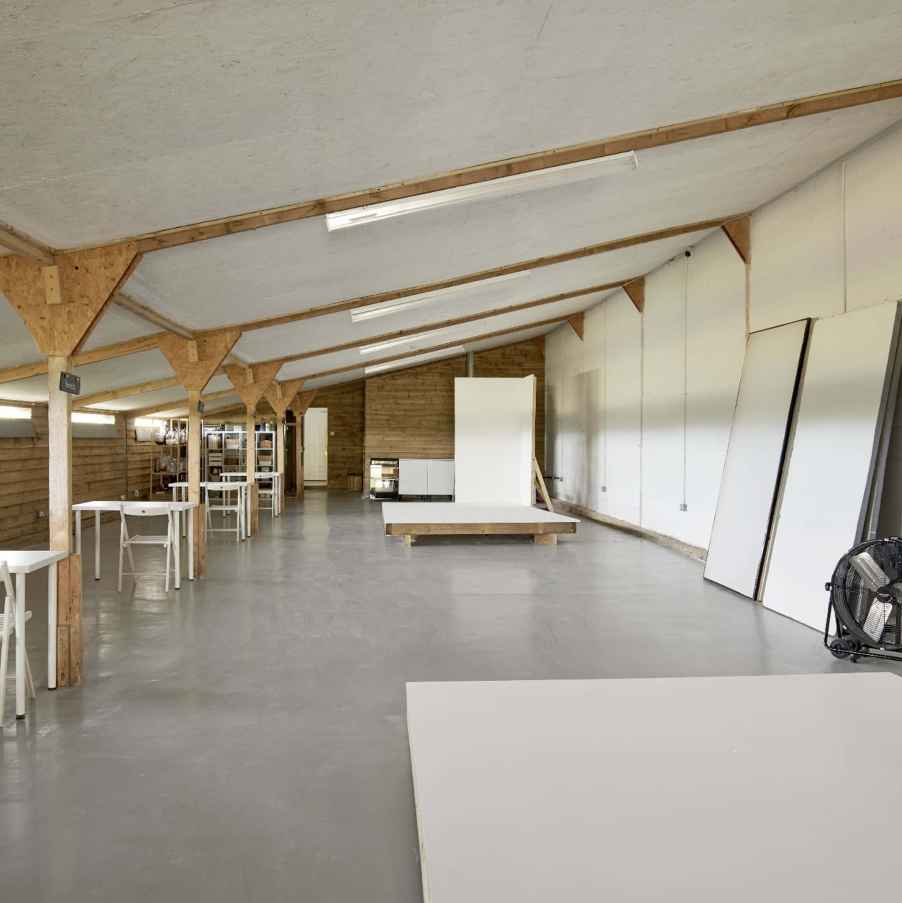 A spacious, minimalist studio with wooden beams, white walls, and empty tables set up for work.