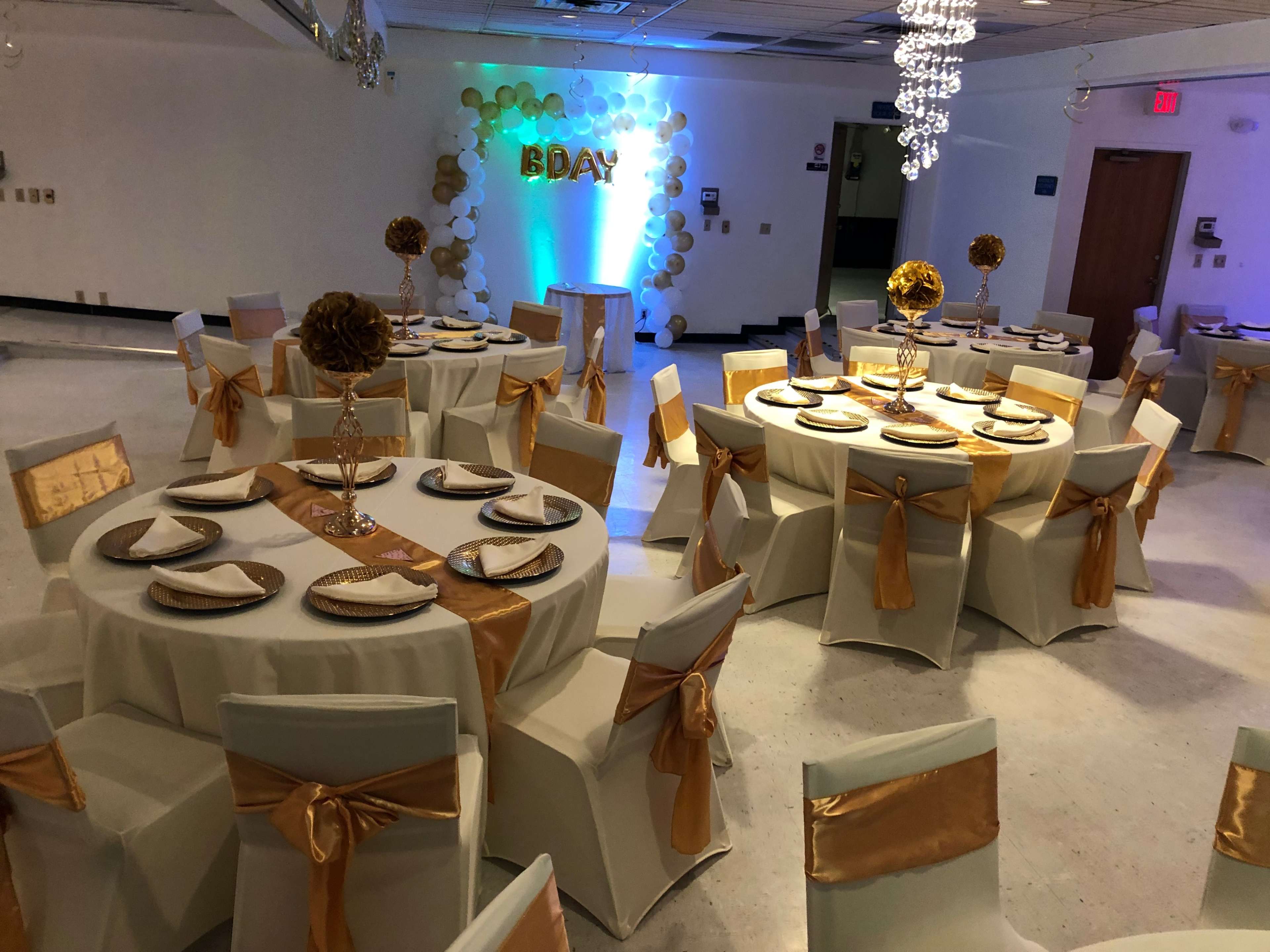 A decorated indoor event space with round tables covered in white tablecloths, gold accents, and a backdrop displaying "BDAY" surrounded by balloons.