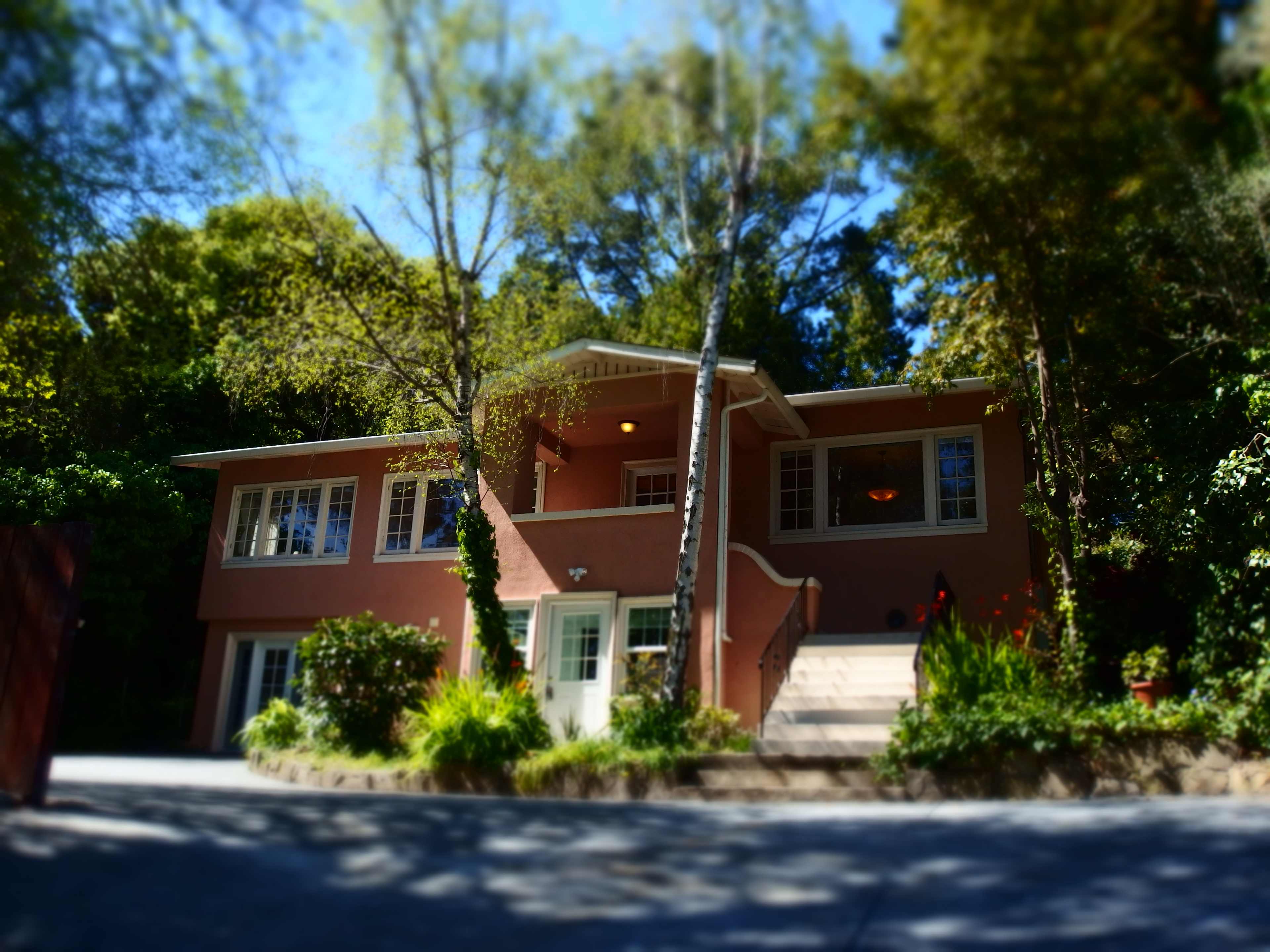 A two-story pink house with a staircase leading to the front door, surrounded by trees and greenery.