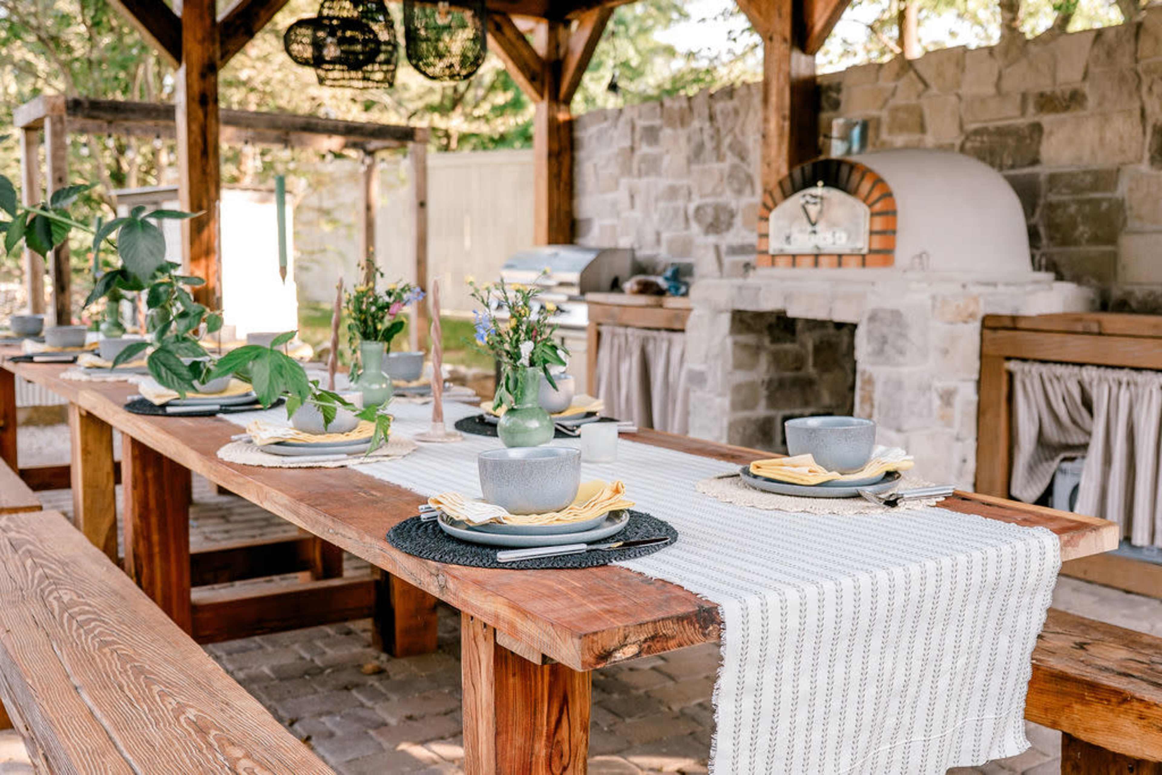 The image shows an outdoor dining area with a large wooden table set for a meal, featuring dishes, utensils, and decorative plants, next to a stone barbecue oven.