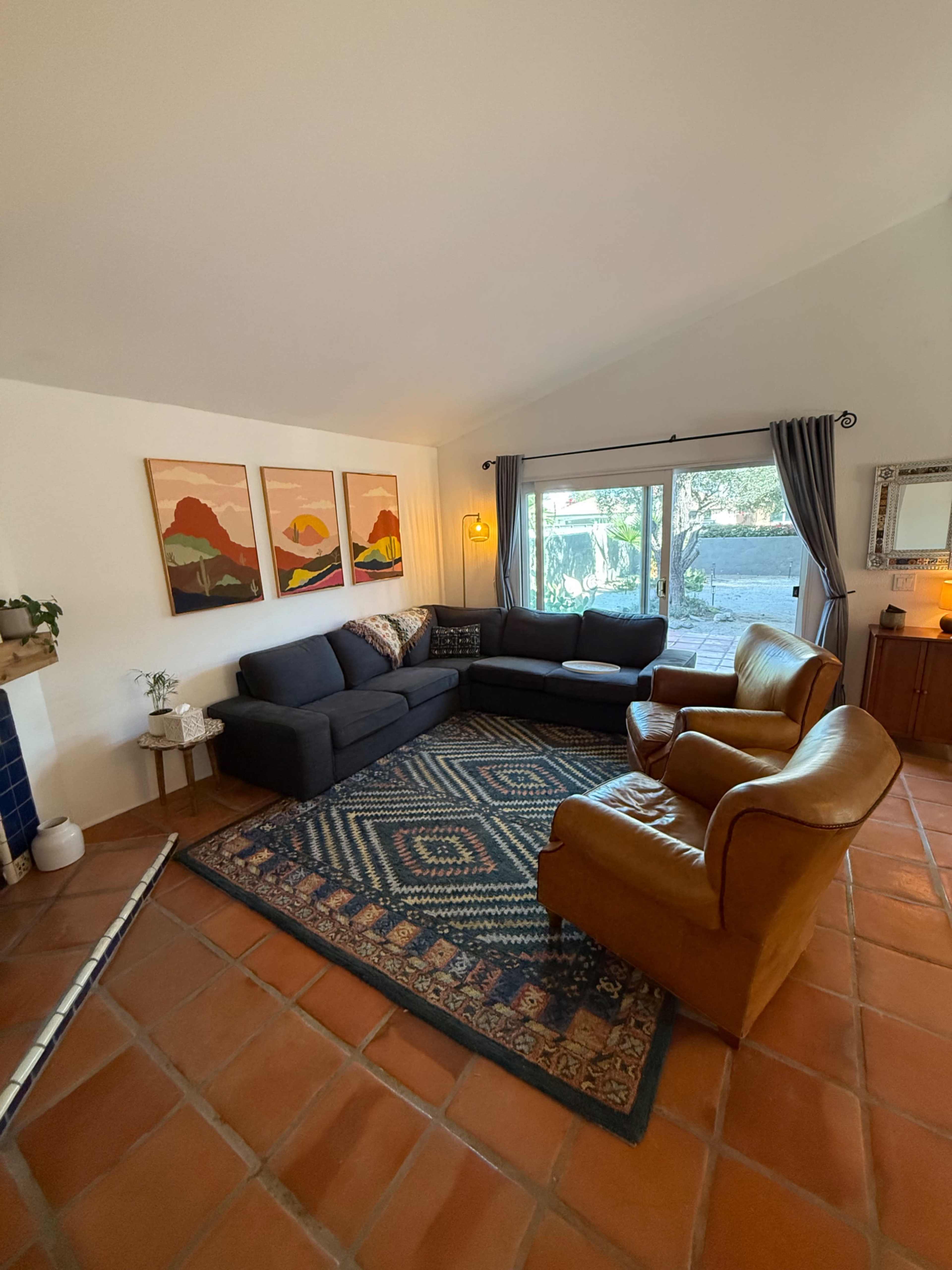 The image shows a living room featuring a sectional sofa, two armchairs, a patterned area rug, and artwork on the walls, all complemented by a large window allowing natural light.