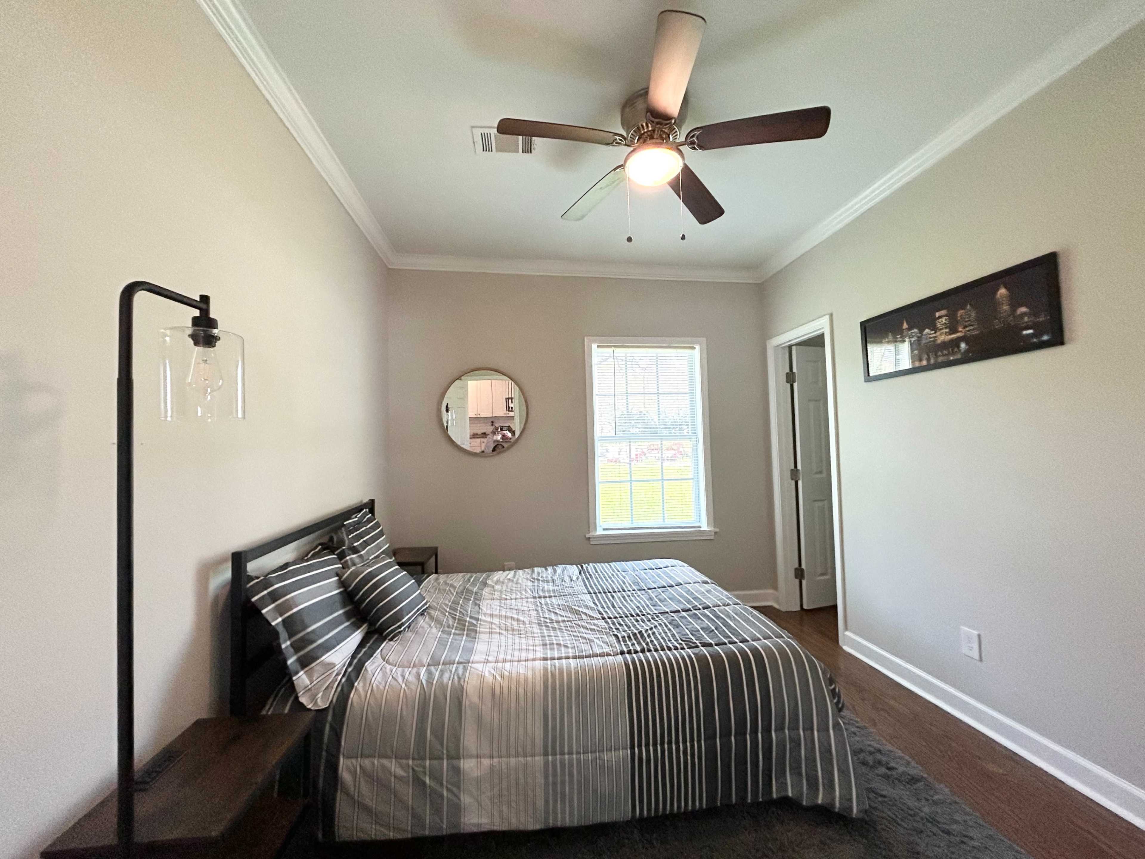 The image shows a simple bedroom featuring a bed with striped bedding, a ceiling fan, a round mirror on the wall, and a bedside lamp beside a nightstand.