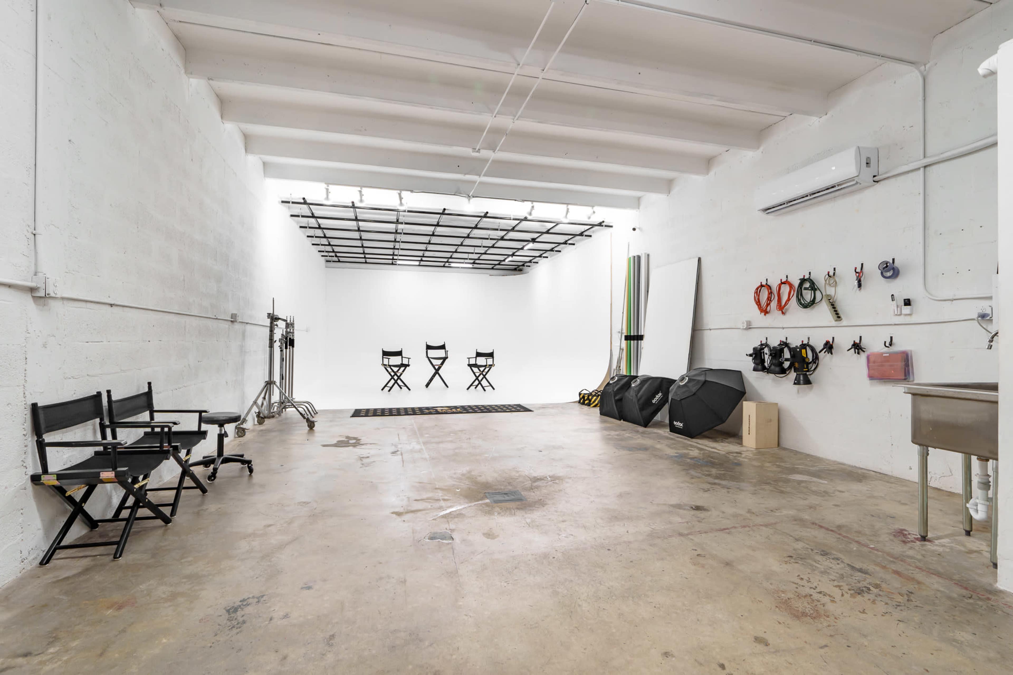 An empty photography studio with three director's chairs, a clean white backdrop, and various equipment on the walls.